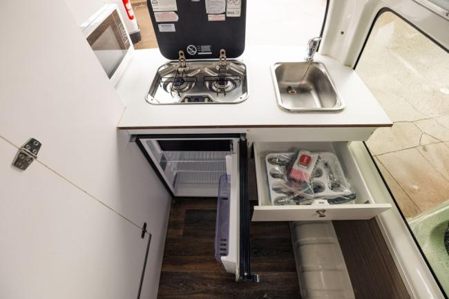 Campervan kitchenette featuring a two-burner gas stove, sink, open mini-fridge, and an open cutlery drawer.