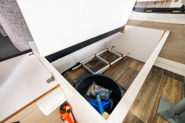 Open under-seat storage in a campervan containing a folded ladder, bucket with tools, and a small fire extinguisher.