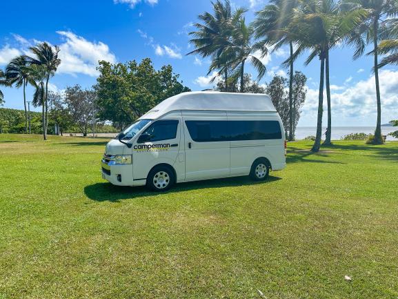 White Camperman high-top campervan parked on grass with palm trees and the ocean in the background.