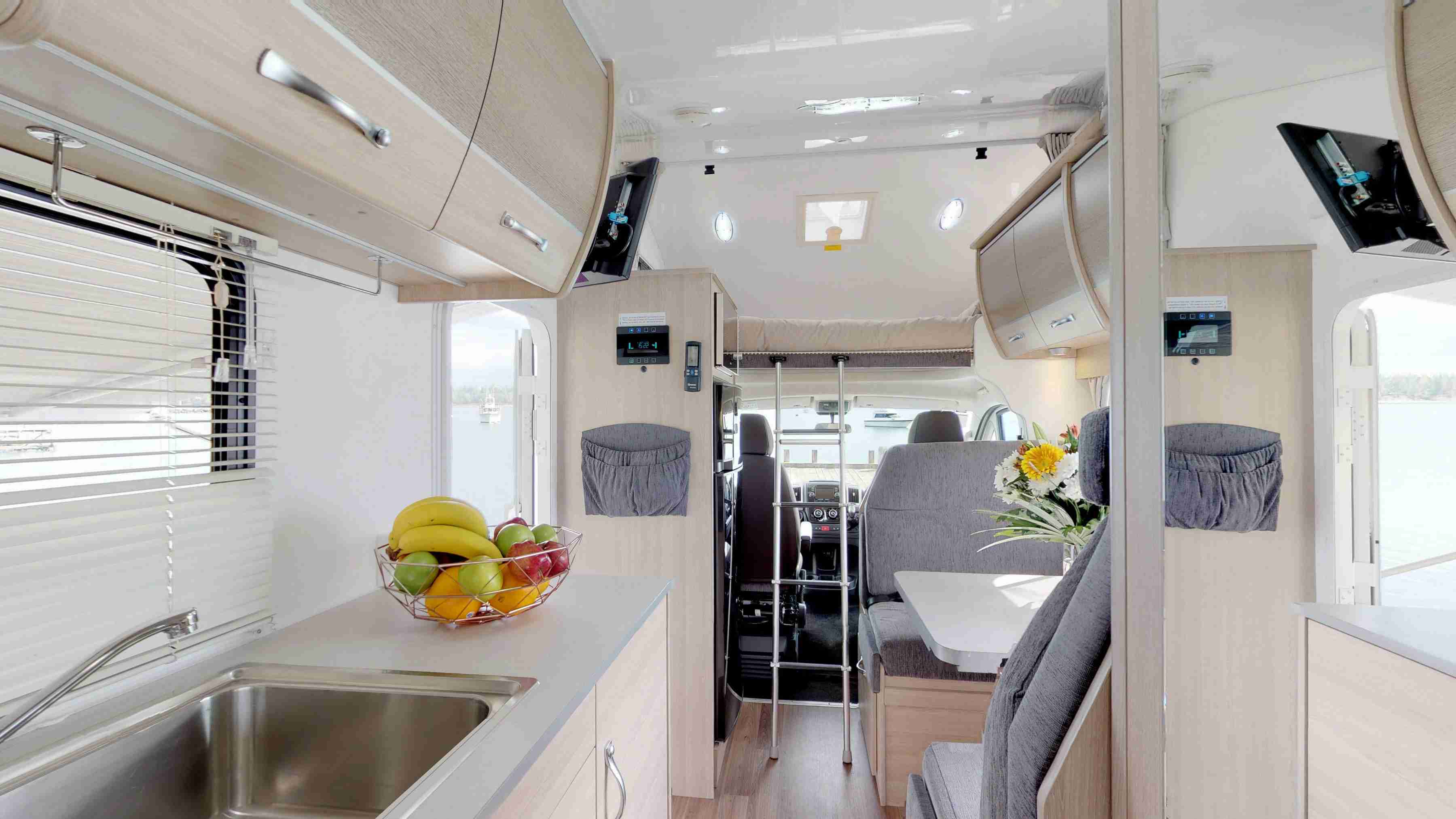 Modern campervan interior with a kitchen sink, fruit bowl, grey dining booth, and an over-cab bed with metal ladder.