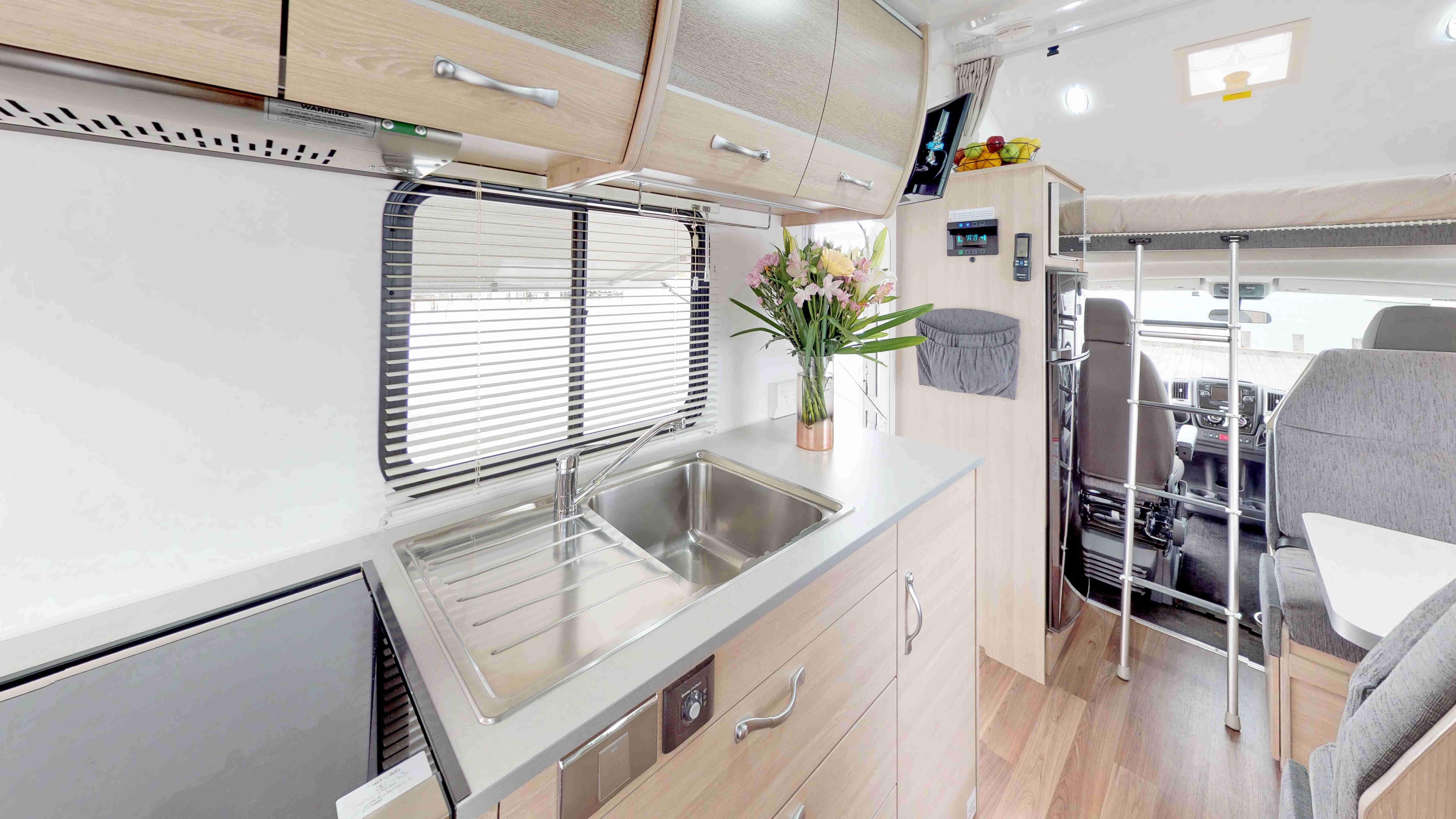 Campervan interior featuring a kitchen with a stainless steel sink, light-toned cabinets, and a ladder to an over-cab bed.