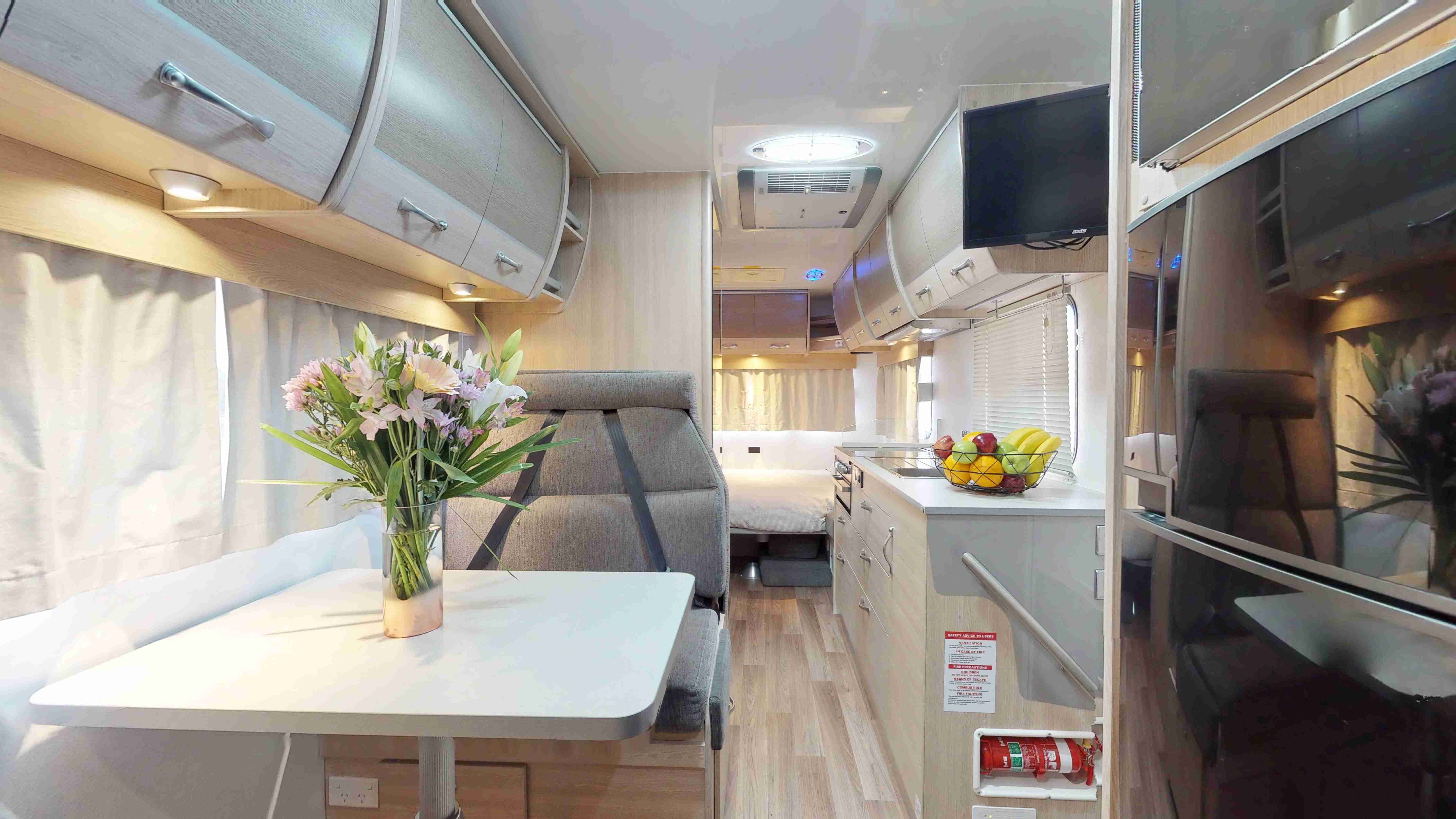 Modern campervan interior with a dining table, grey fabric seats, kitchenette, and a double bed in the background.