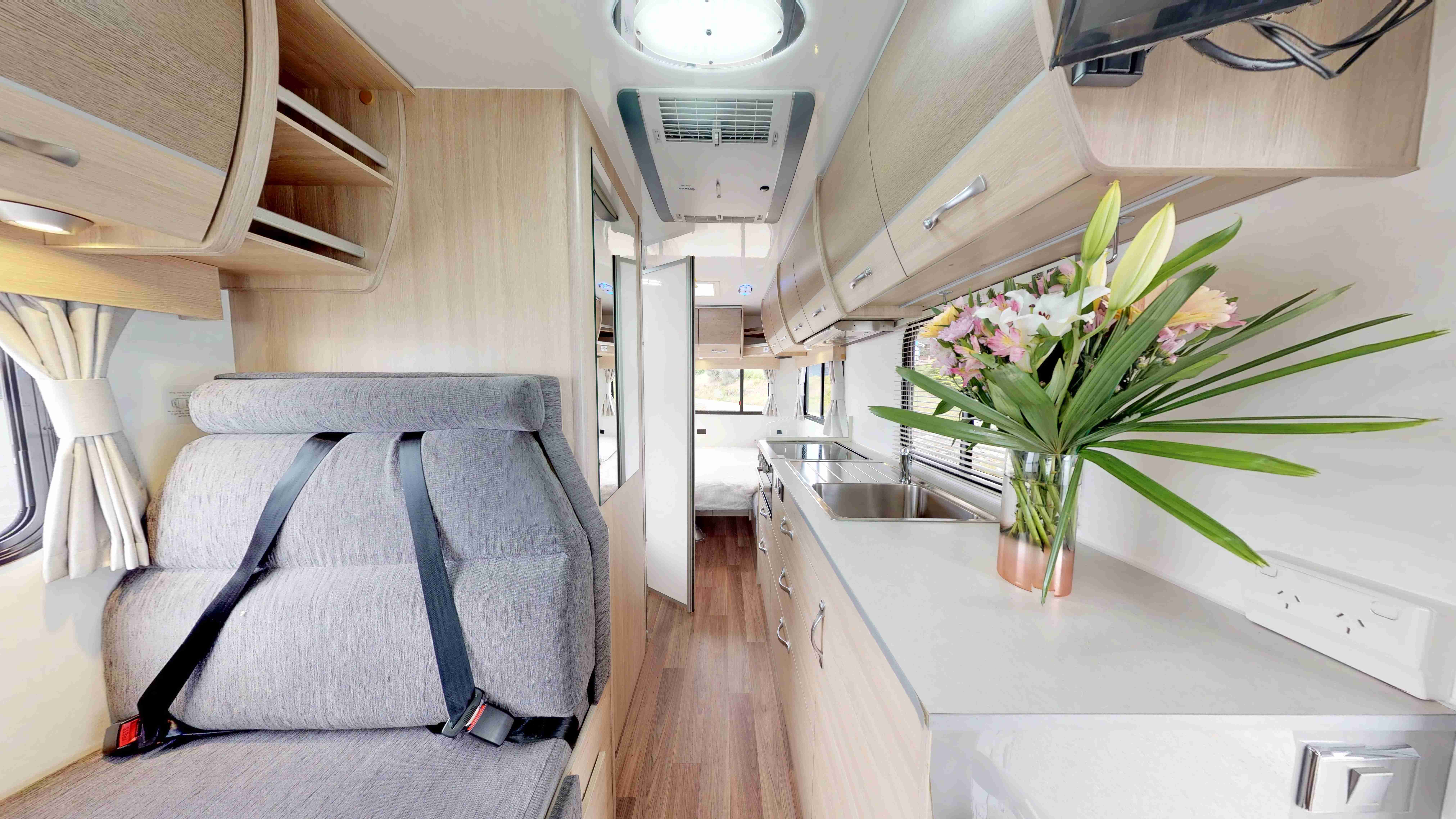 Campervan interior featuring a grey seat with seatbelts, kitchen counter with sink, and hallway leading to a rear bed.