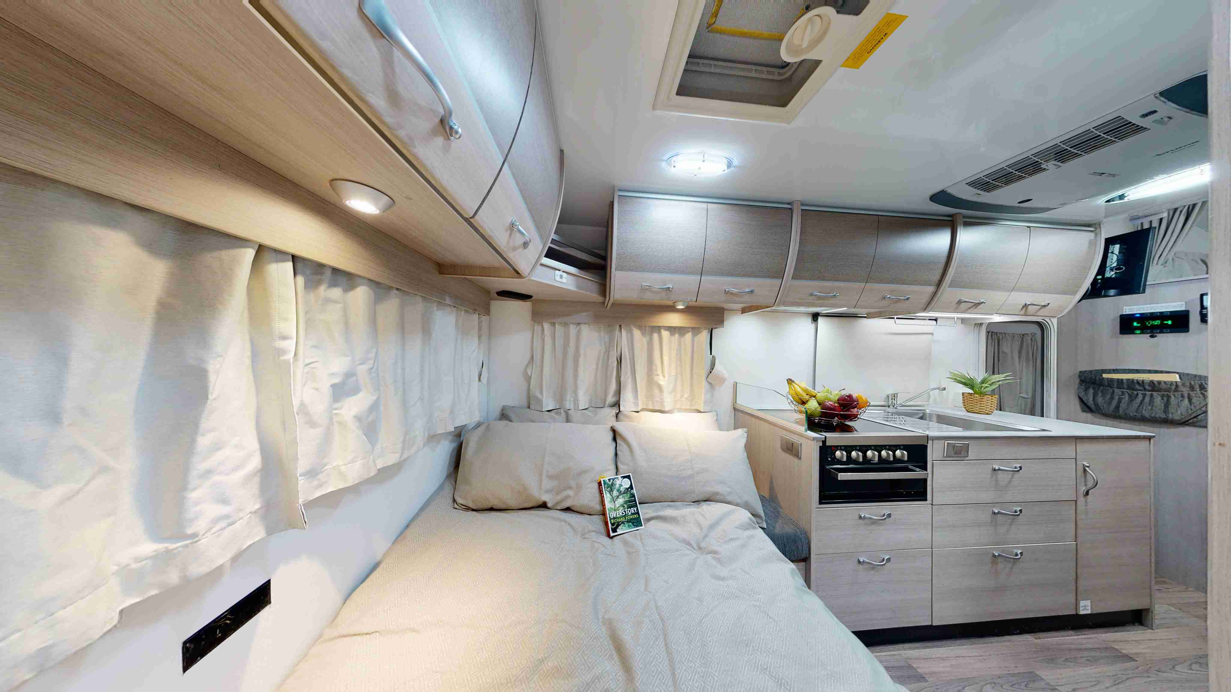 Campervan interior featuring a made bed, overhead storage, and a compact kitchen with light timber cabinets.