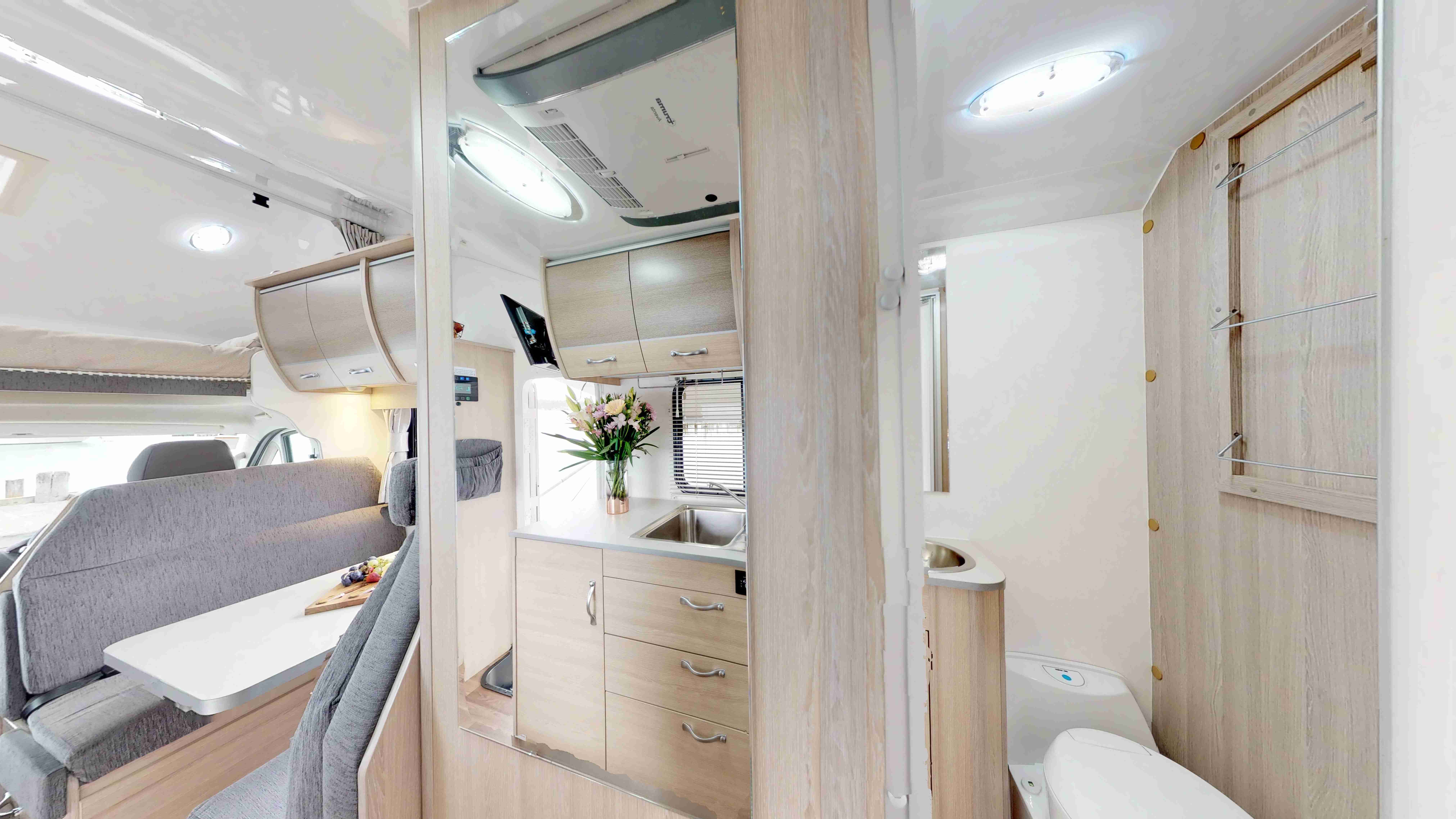 Campervan interior featuring a grey dinette, a mirrored kitchen with a sink, and a compact bathroom with a toilet.