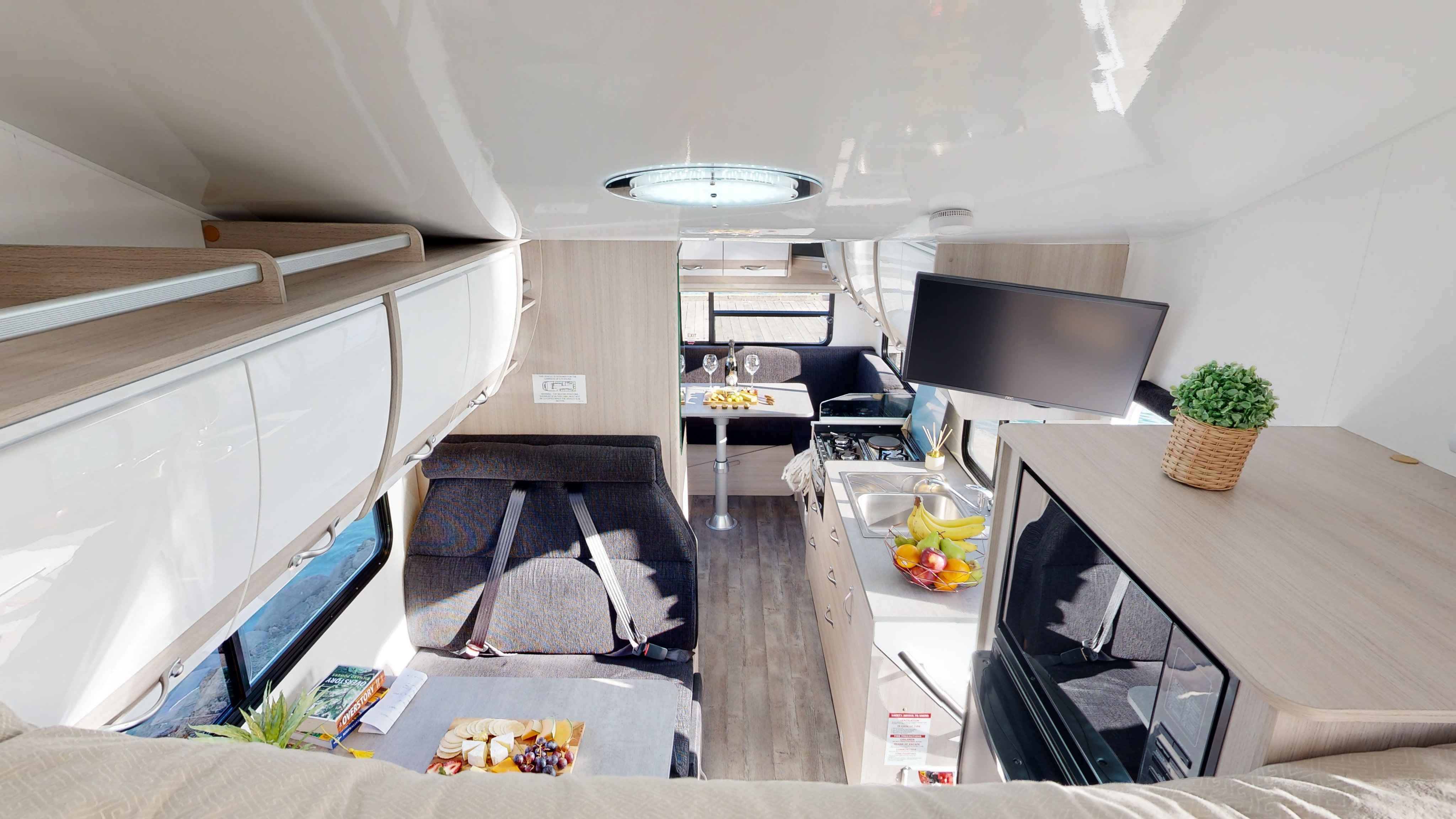 Campervan interior featuring light timber-look cabinets, a grey dinette, compact kitchen, and rear dining area.