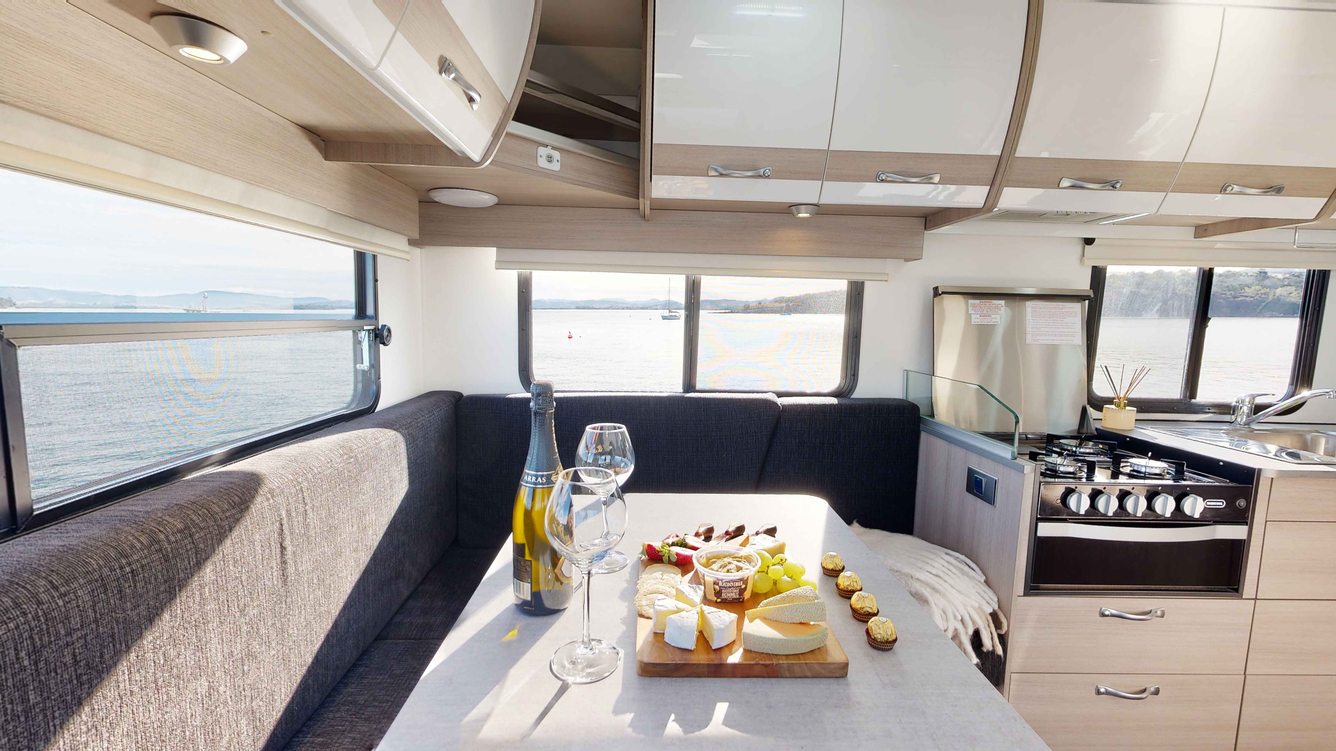 Campervan interior showing a dining table with sparkling wine, cheese platter, and glasses next to a window with water views.