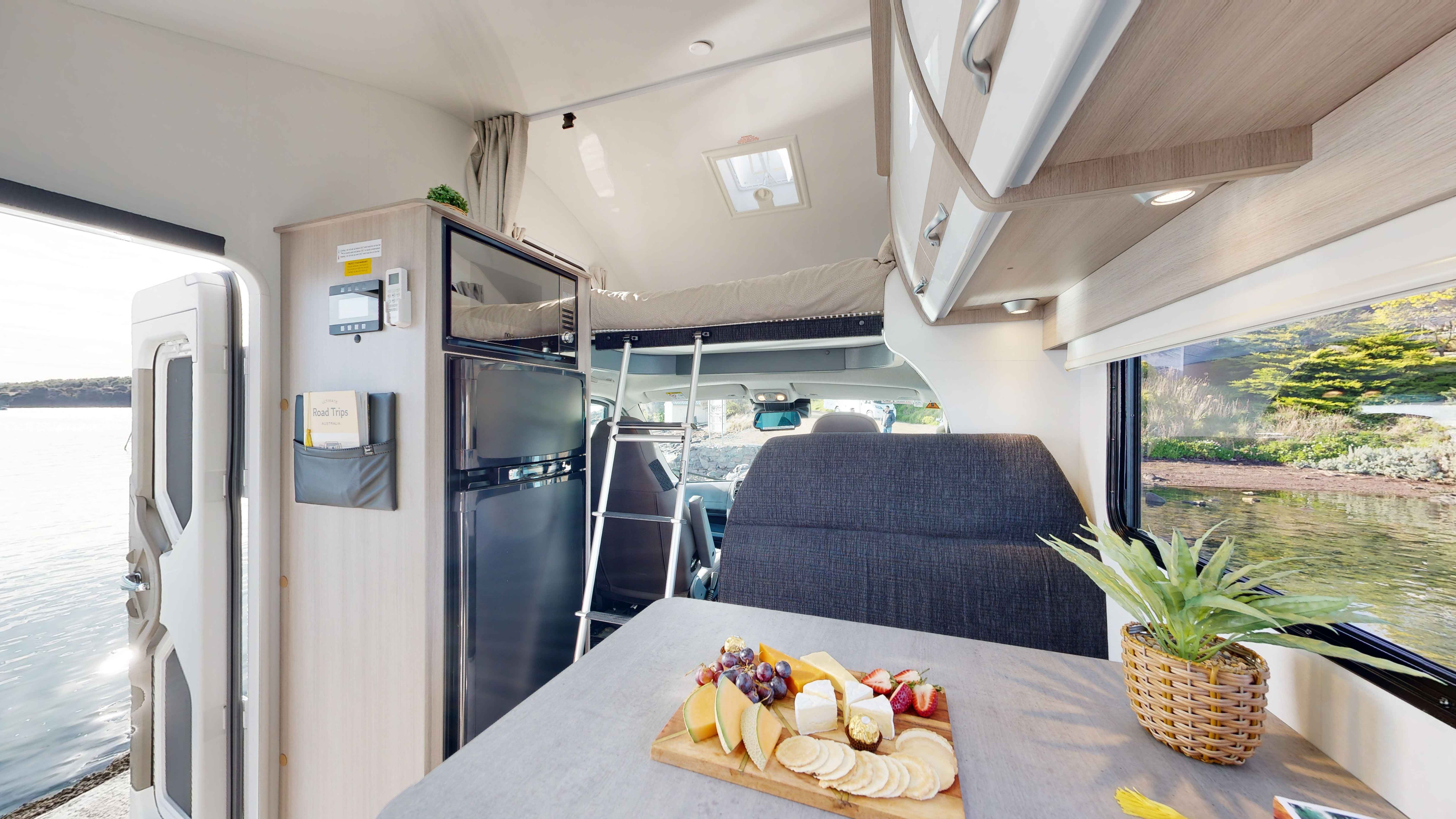 Modern campervan interior with a cheese platter on the table, a loft bed ladder, and scenic outdoor views.
