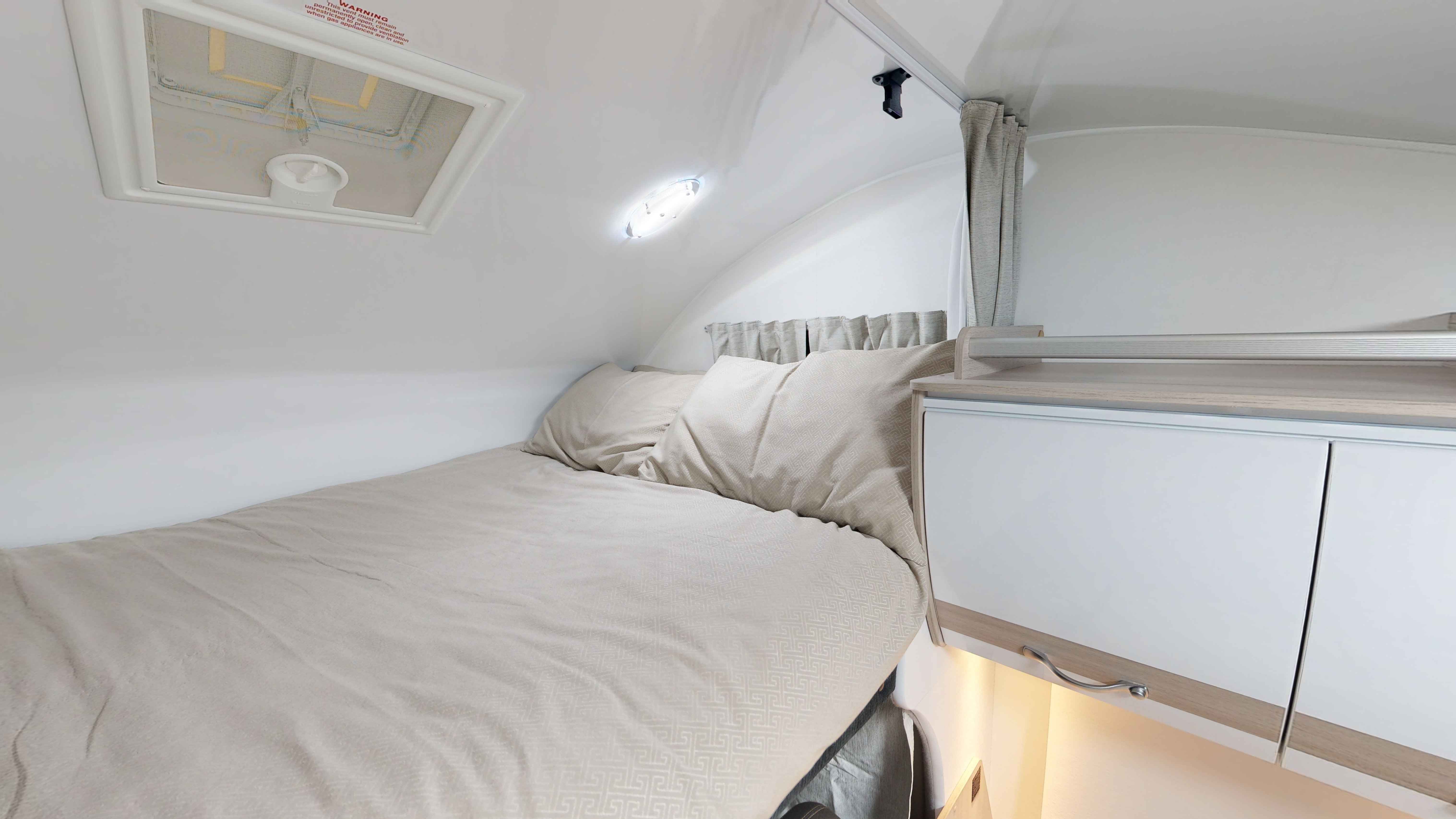 Compact campervan interior featuring a bed with beige bedding, curved white walls, a roof vent, and white cabinets.