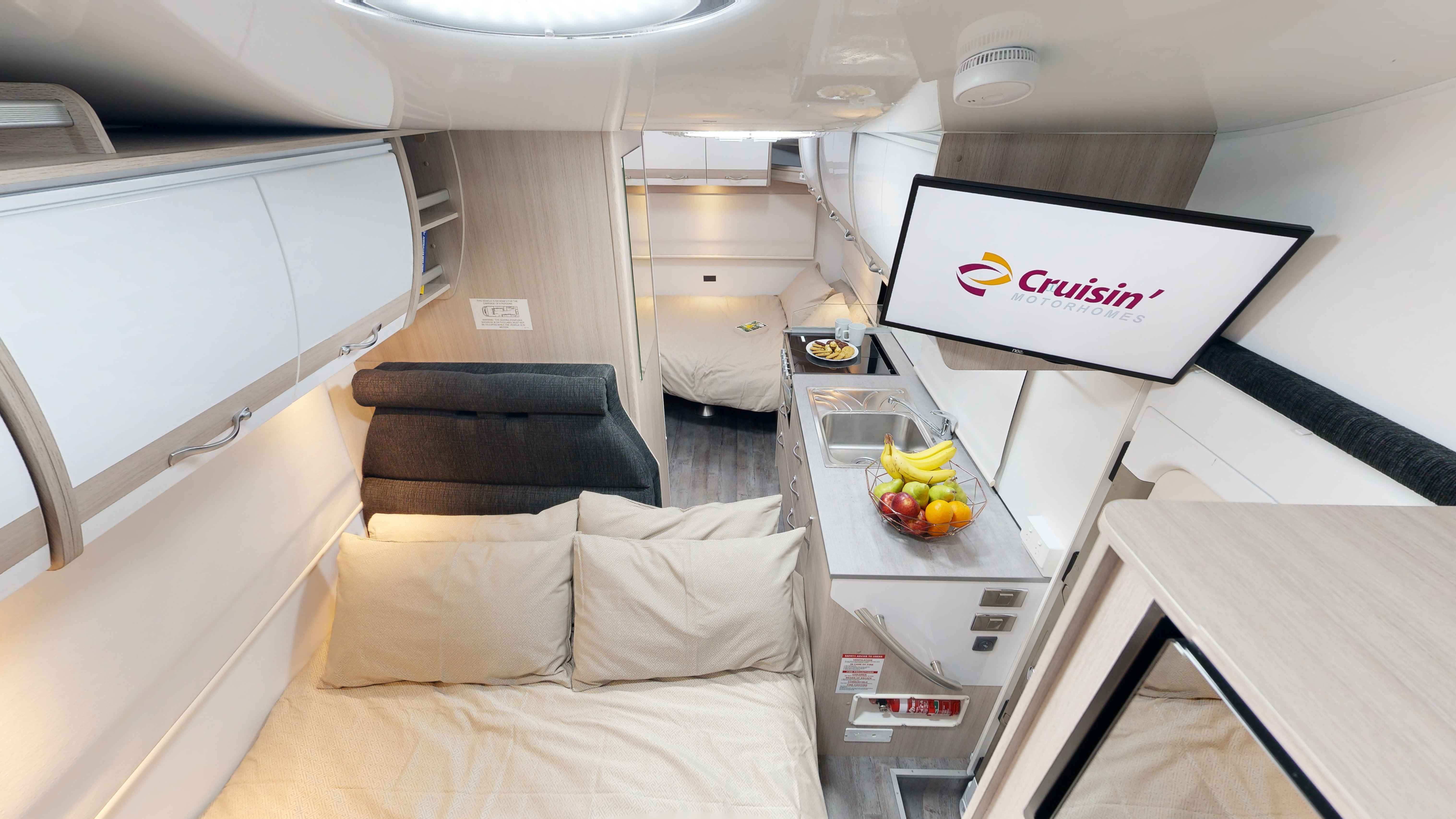 Interior of a campervan showing two beds, a small kitchen with fruit, and a TV displaying the Cruisin' Motorhomes logo.