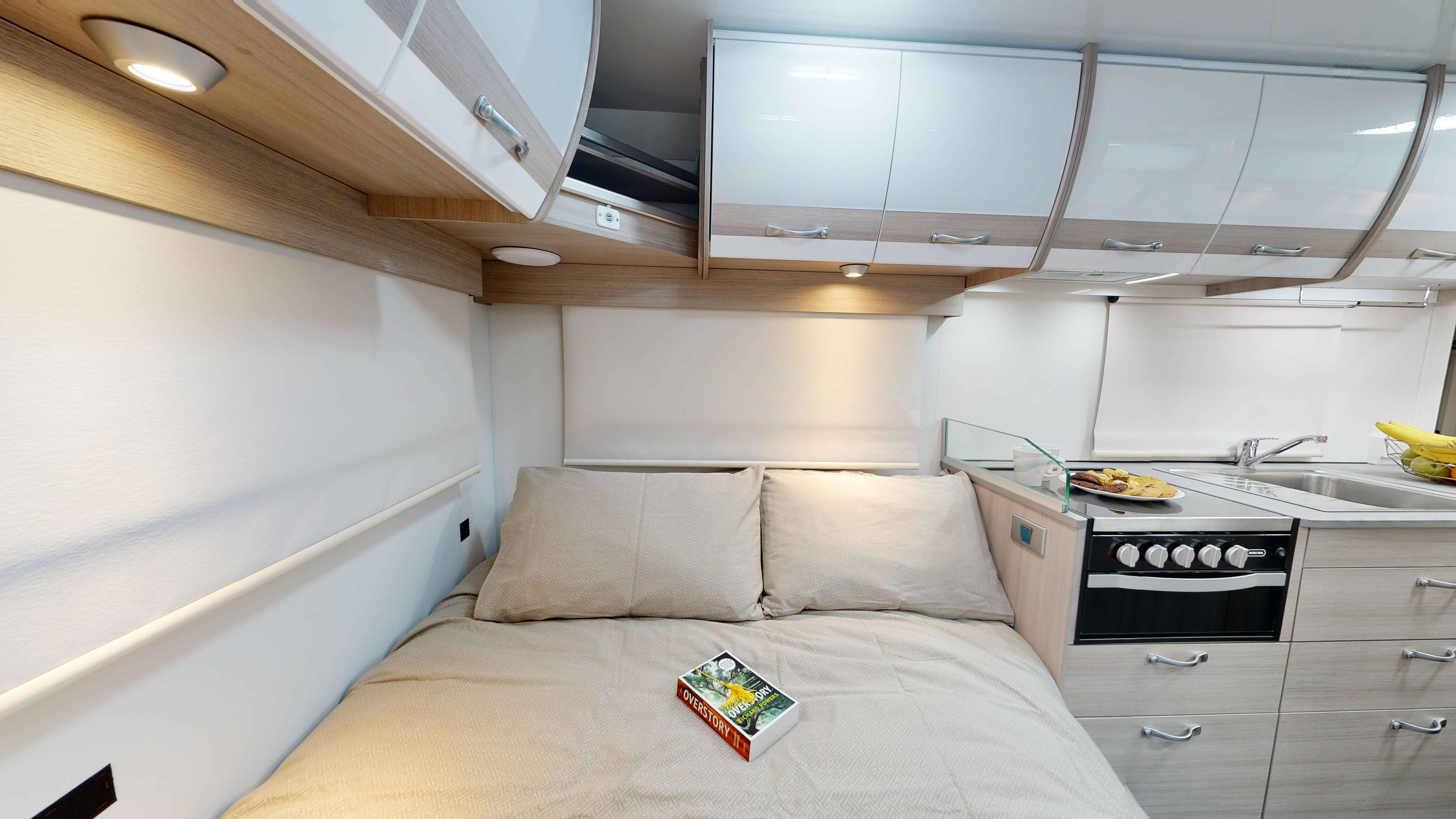 Campervan interior showing a made double bed with a book on top, overhead cabinets, and a compact kitchenette.