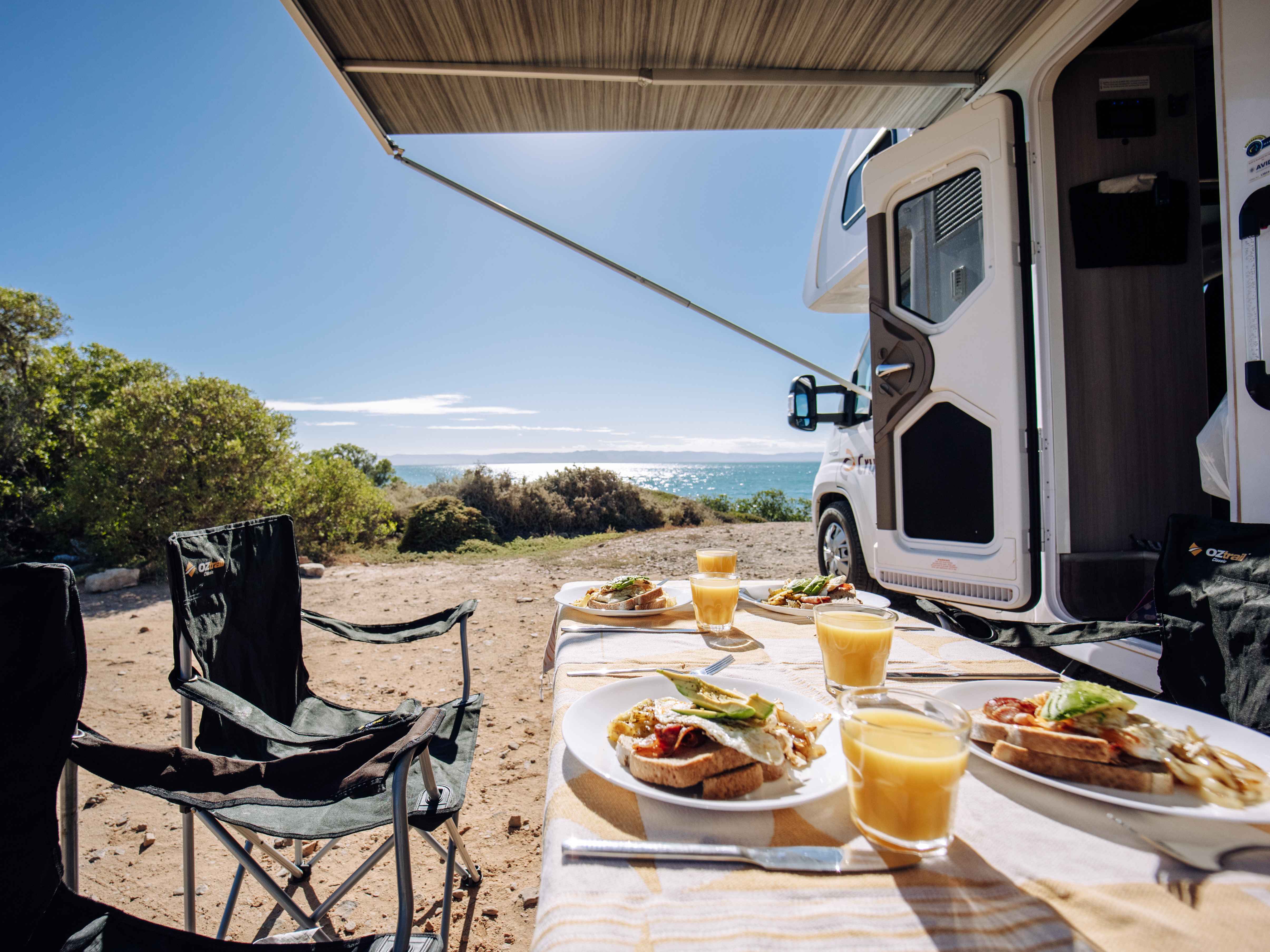Breakfast served on an outdoor table beside a campervan under its awning, overlooking a coastal ocean view.