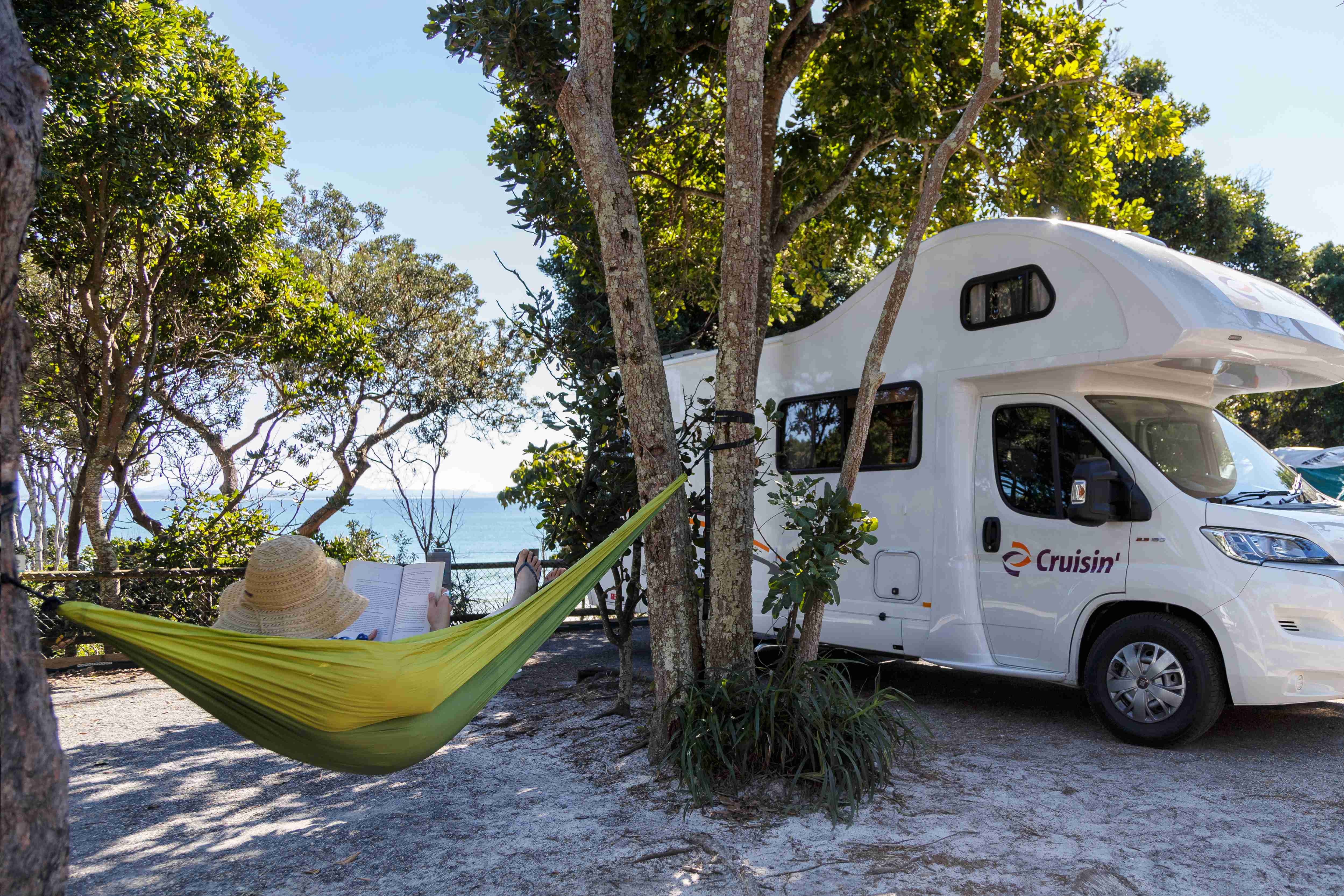 Person in a straw hat reading in a hammock next to a white Cruisin' campervan parked under trees near the sea.