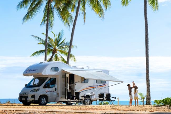 White campervan with extended awning parked under palm trees by the sea, with a family standing nearby.