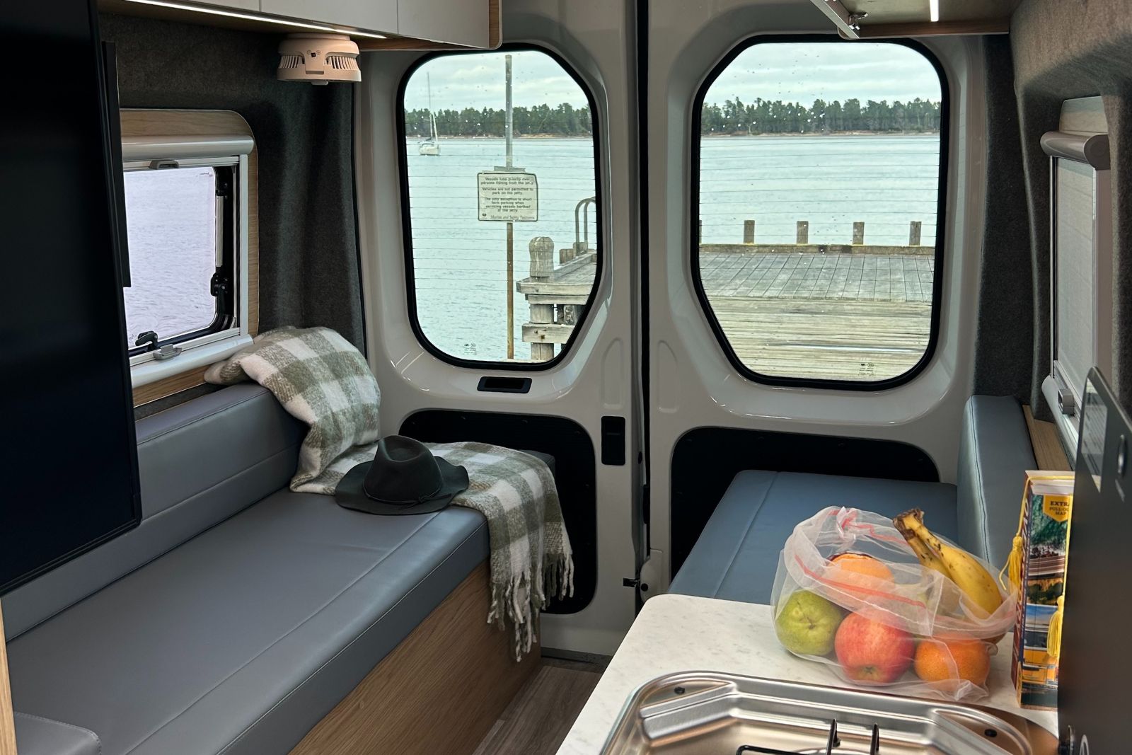 Campervan interior looking through rear door windows at a jetty and water, with grey bench seats and a bag of fruit.