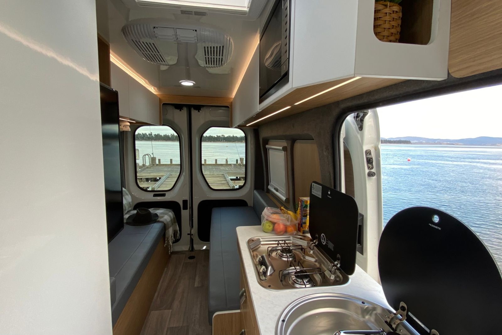 Interior of a modern campervan with a kitchenette, bench seats, and open doors looking out onto a jetty and the sea.