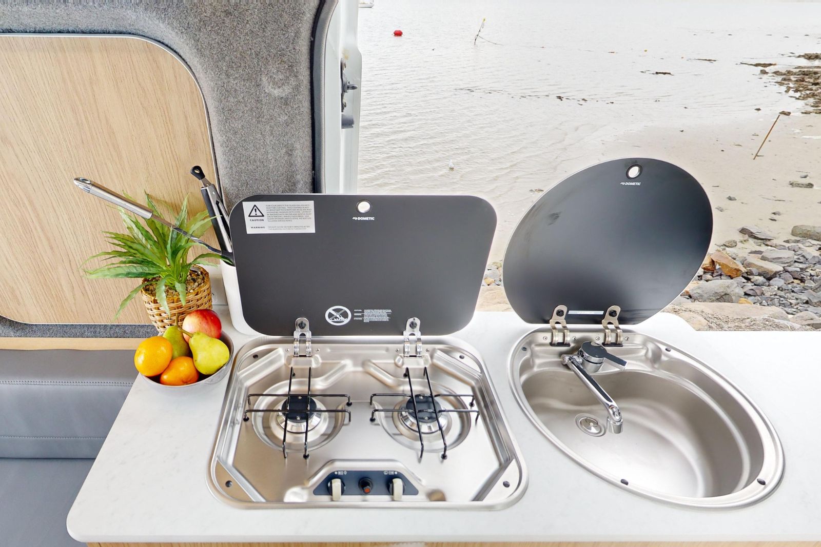 Campervan kitchenette with a two-burner gas stove, sink, and fruit bowl, overlooking a beach.