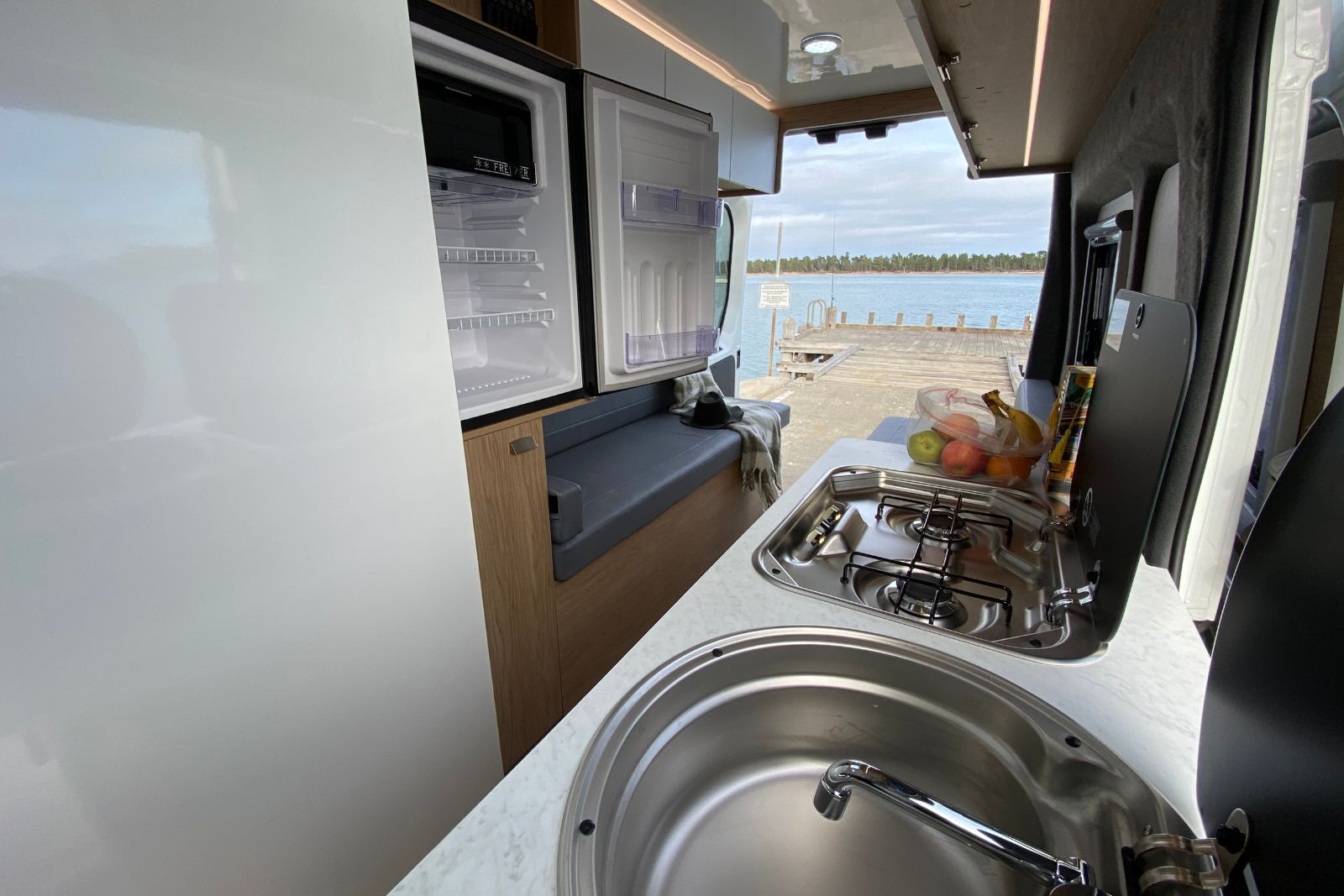 Campervan kitchen with a sink, gas stove, open fridge, and grey bench seat looking out to a timber jetty over water.