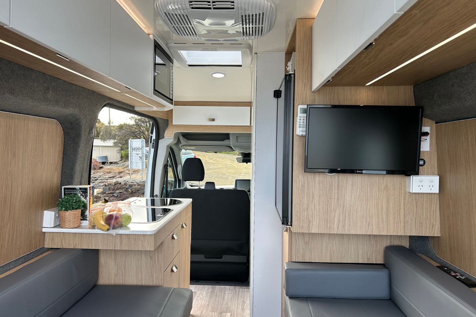 Campervan interior featuring grey leather seats, timber-finish cabinets, a kitchenette, and a wall-mounted TV.