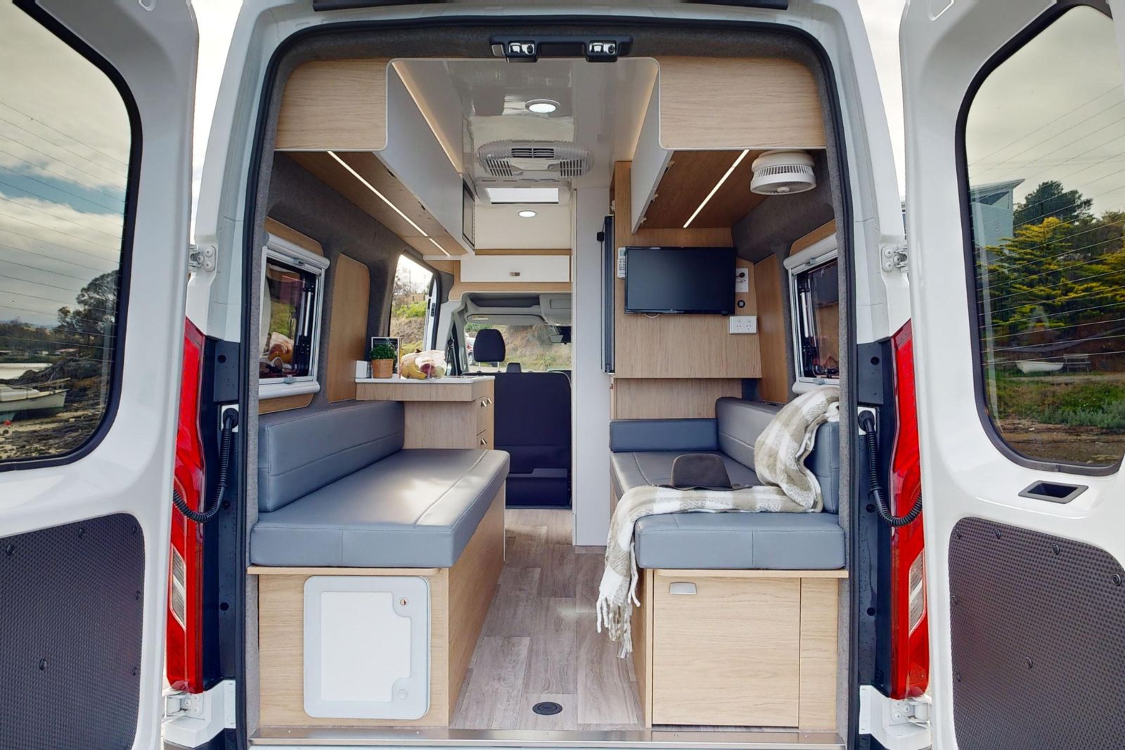 Campervan interior with twin grey bench seats, light timber cabinetry, and a central aisle leading to the front cab.