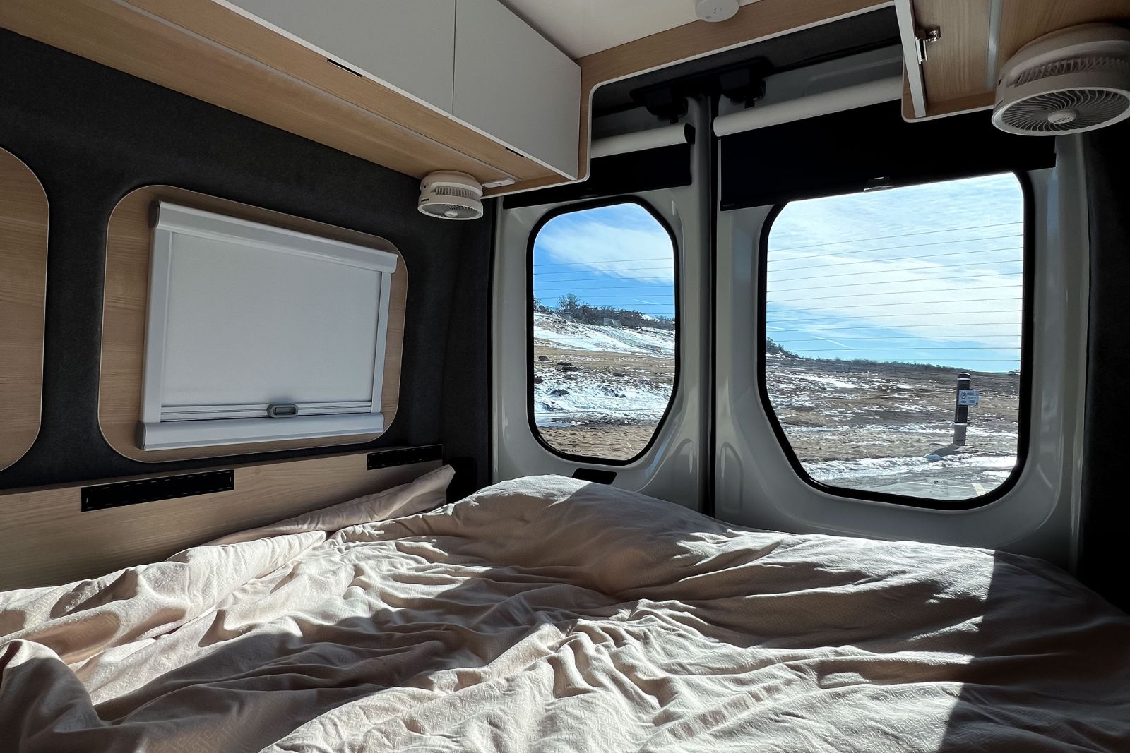 Campervan interior featuring an unmade bed with views of a snowy landscape through two rear door windows.
