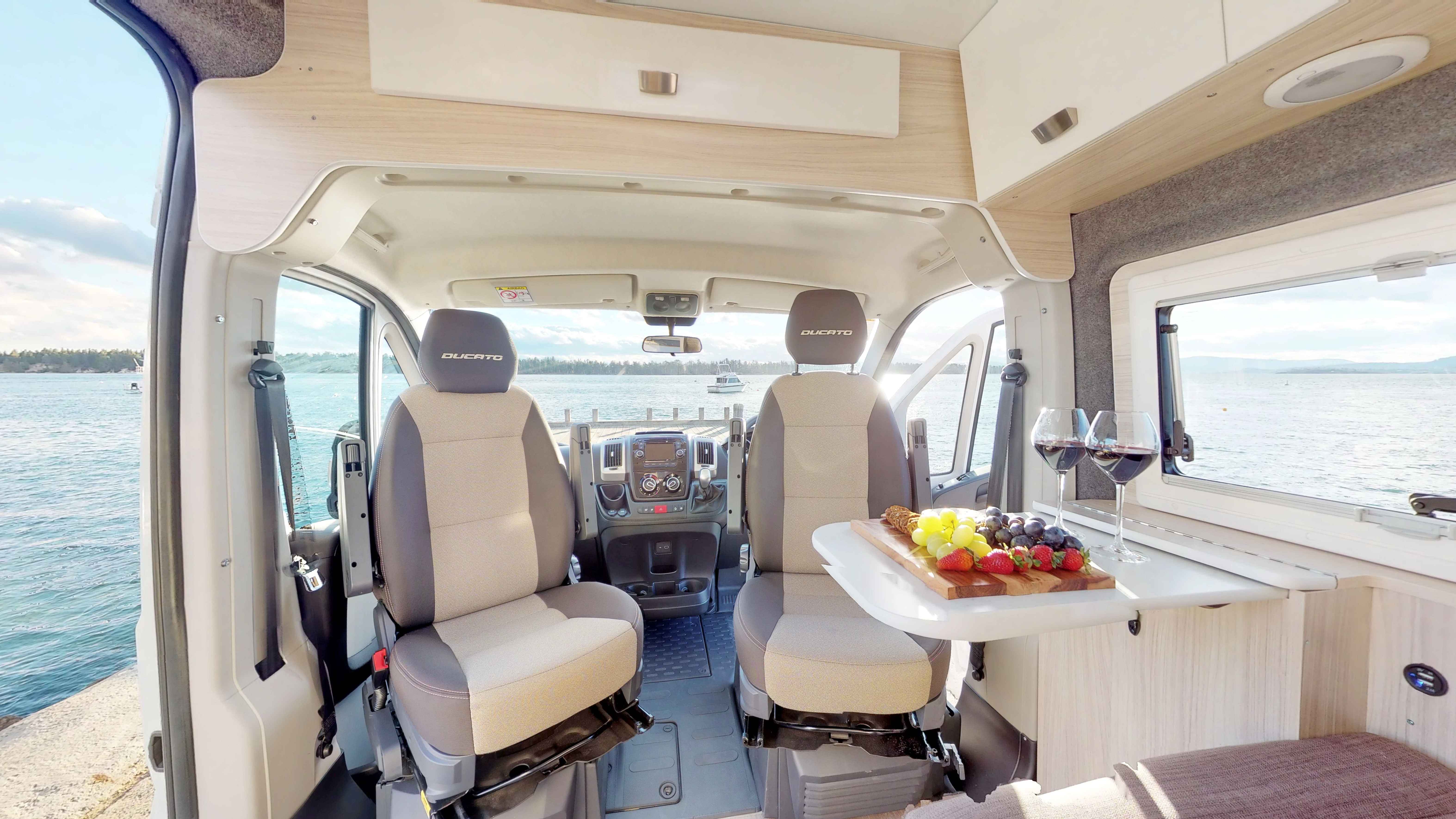 Campervan interior with swivel seats facing a table with red wine and fruit, overlooking a coastal view.