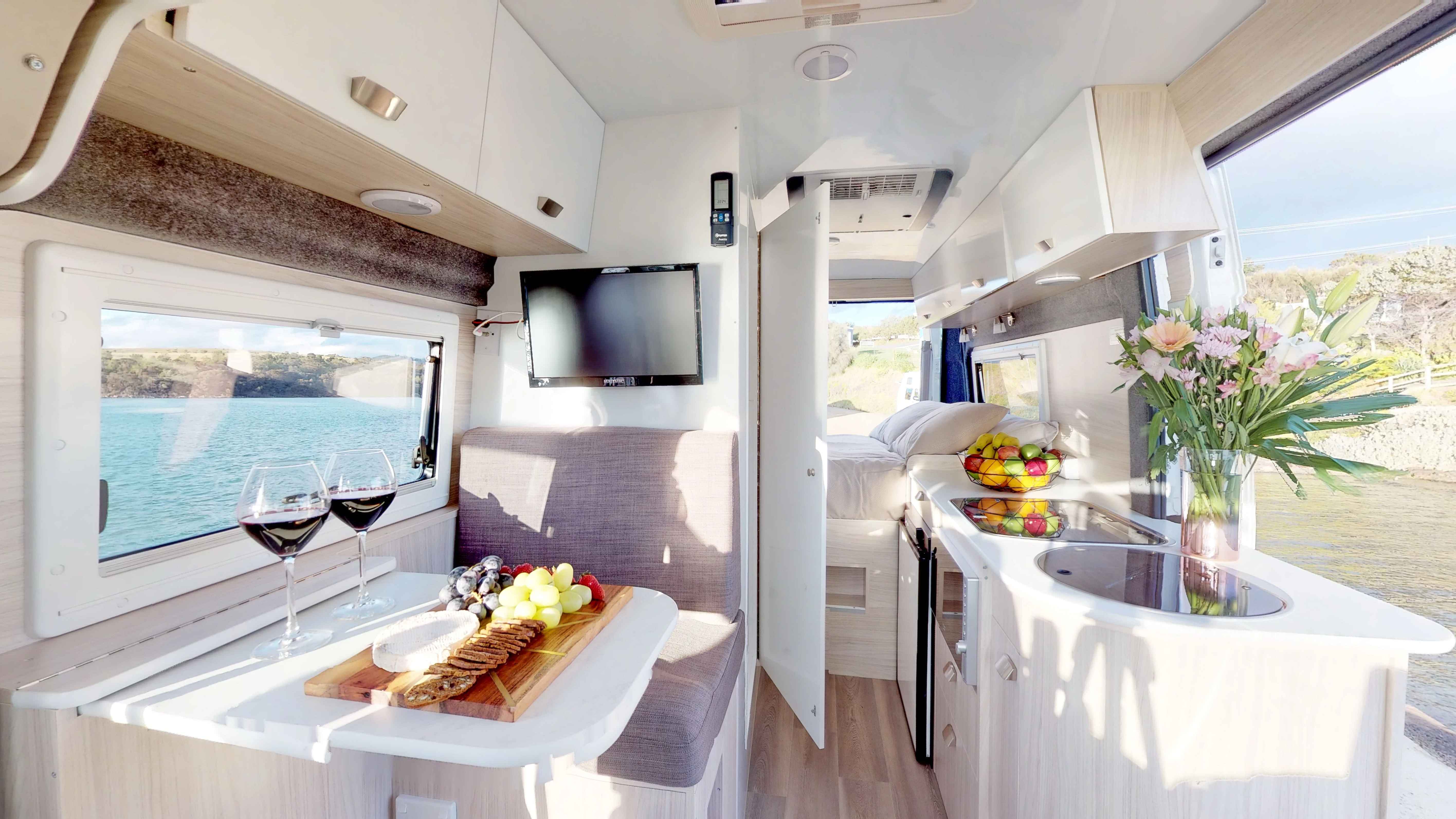 Campervan interior featuring a dining table with wine and cheese, a side kitchenette, and a bed in the background.