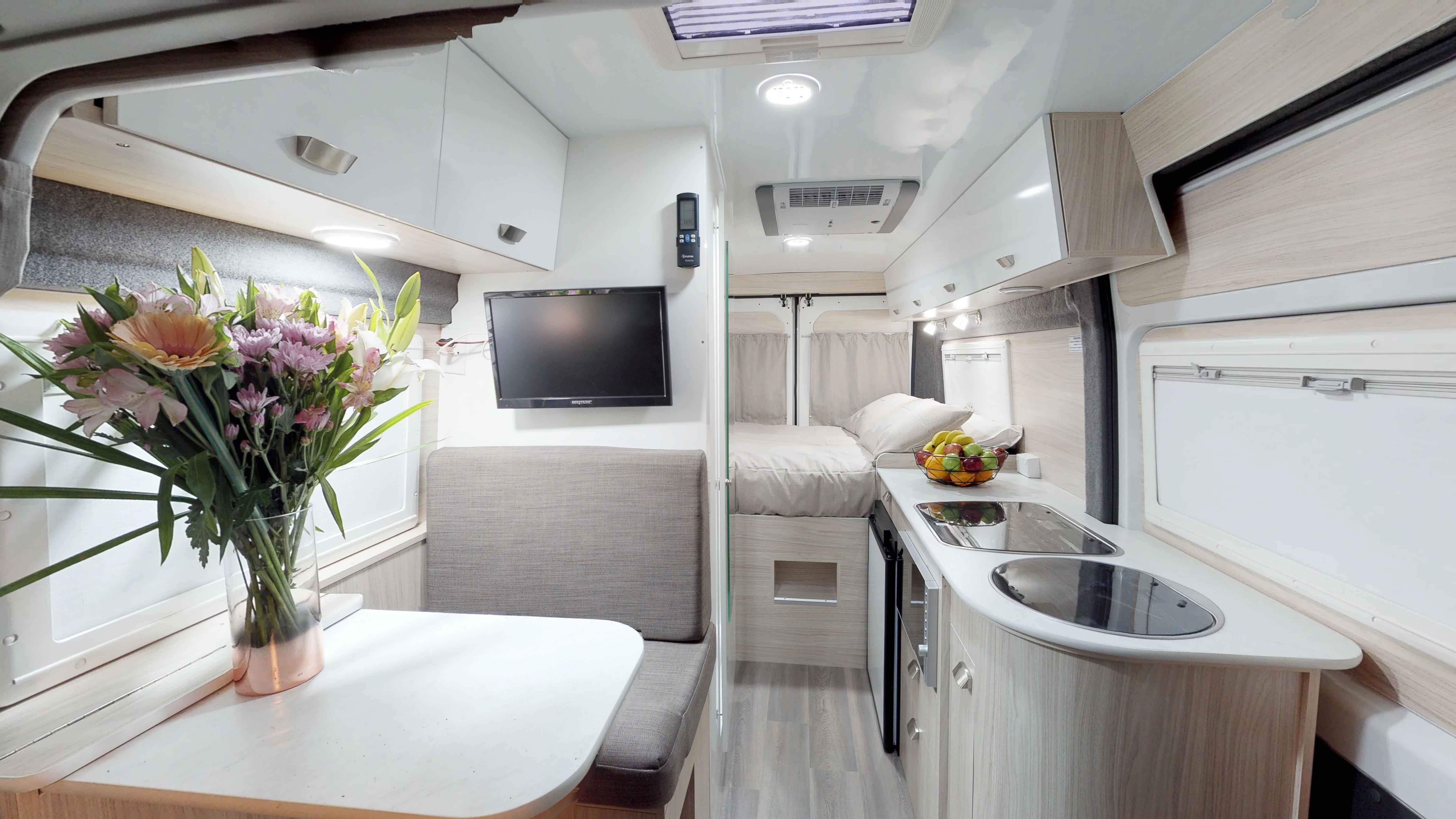 Compact campervan interior featuring a dining table with flowers, a grey booth seat, kitchenette, and a rear double bed.