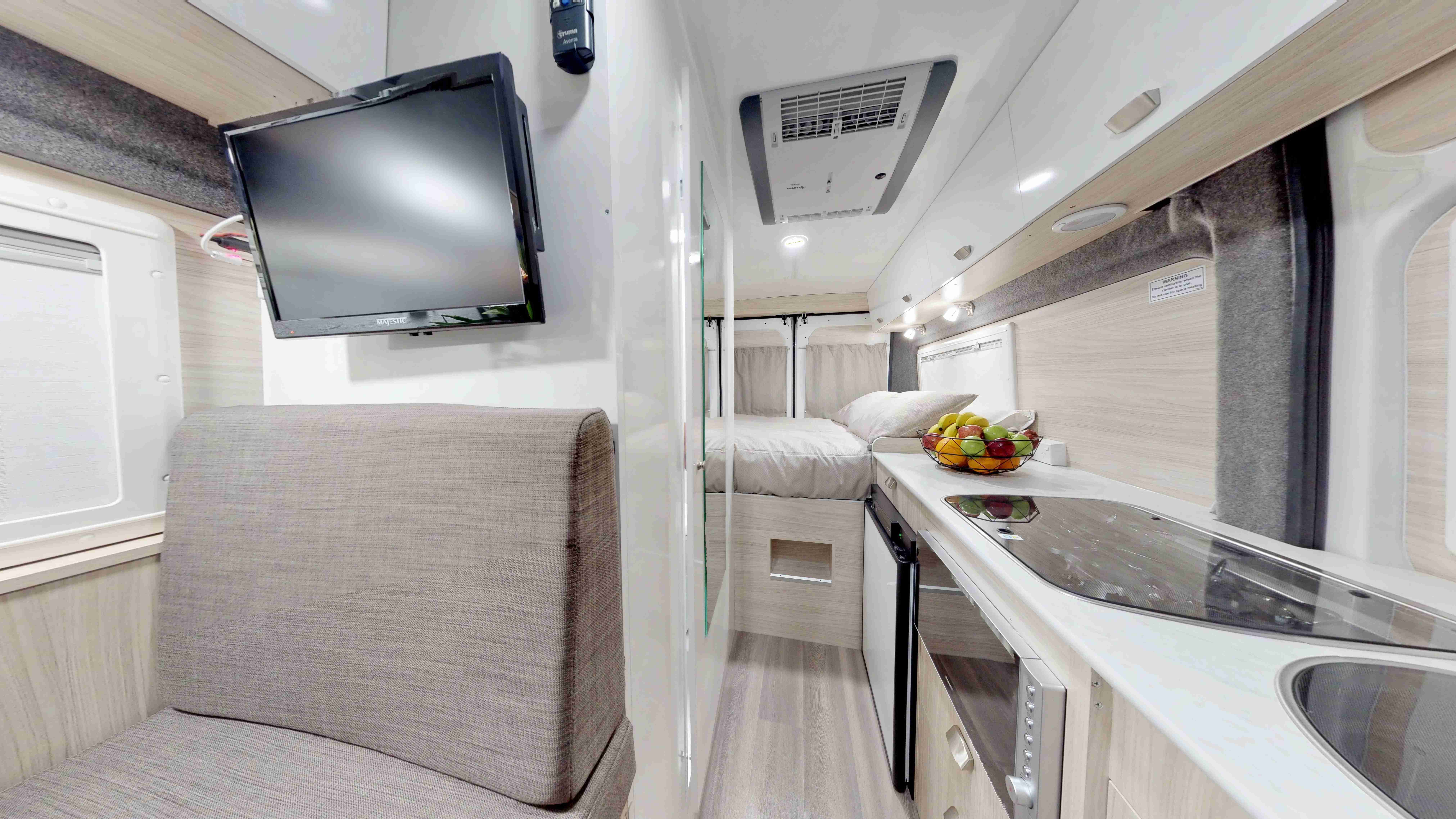 Modern campervan interior with a grey fabric seat, wall-mounted TV, light timber-look kitchen counter, and rear bed.