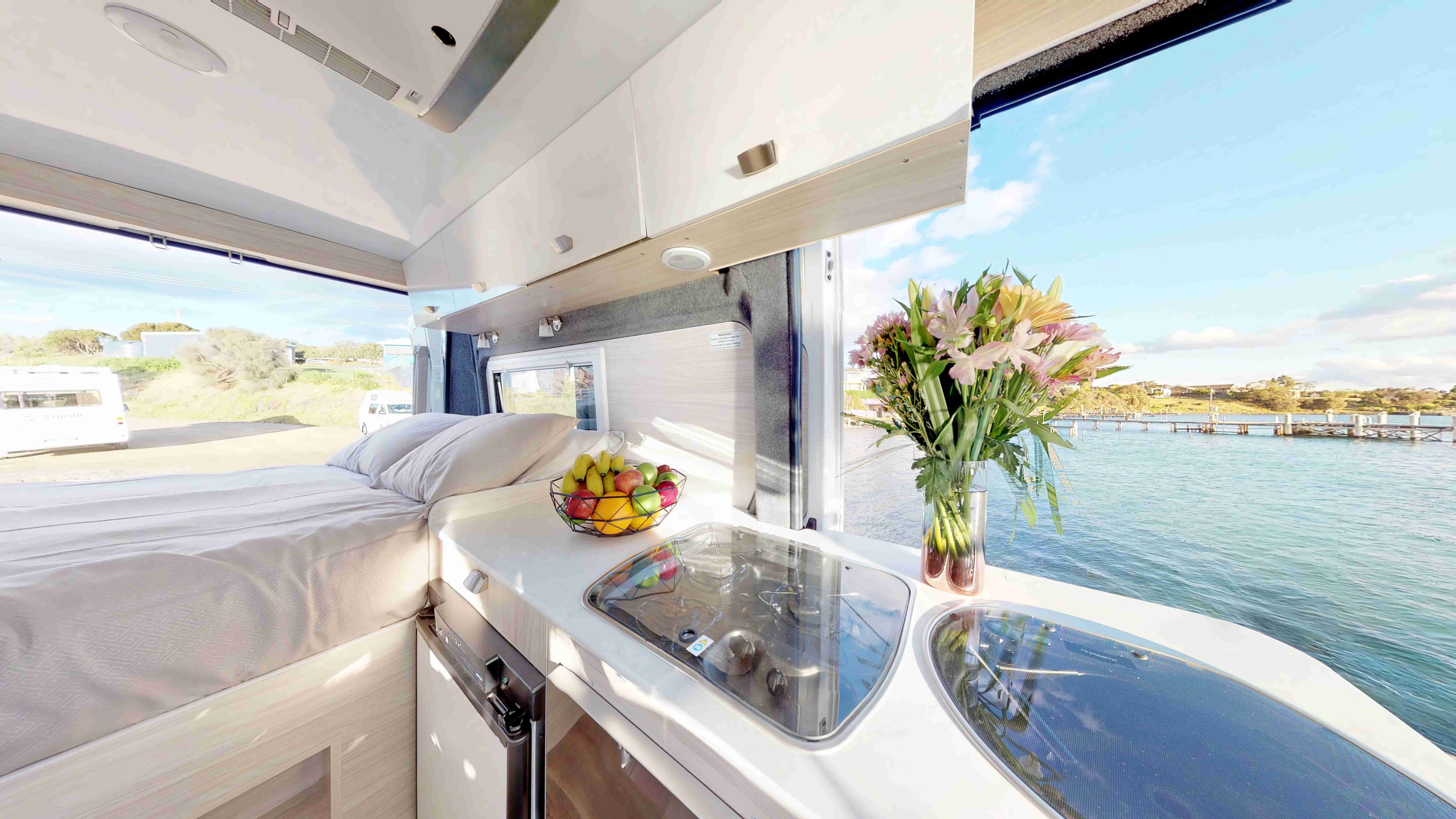 Campervan interior showing a bed and kitchen counter with fruit and flowers, looking out to a sunny harbour.
