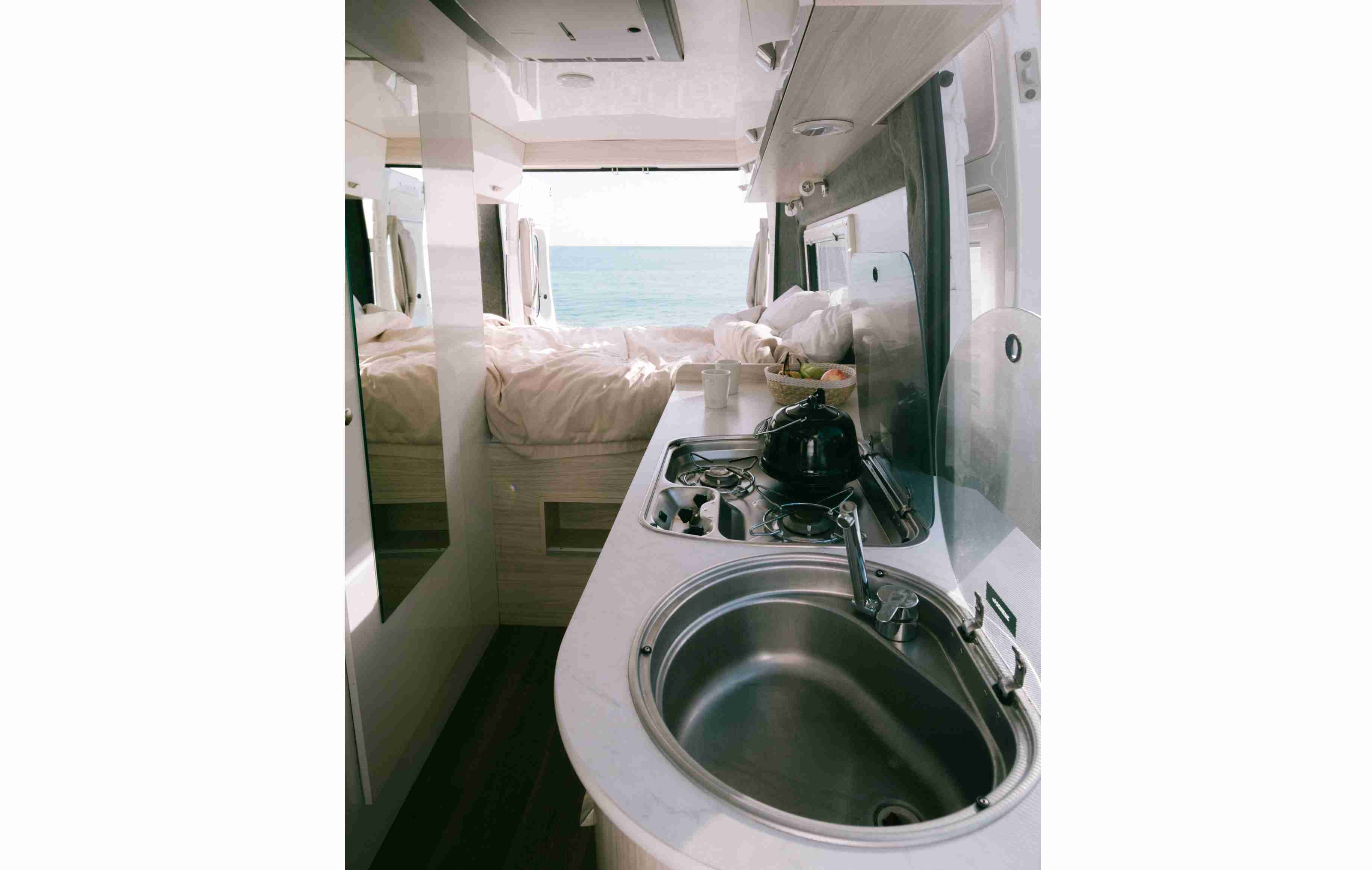 Campervan interior looking past a kitchen sink and stove to a double bed with open doors overlooking a calm ocean.