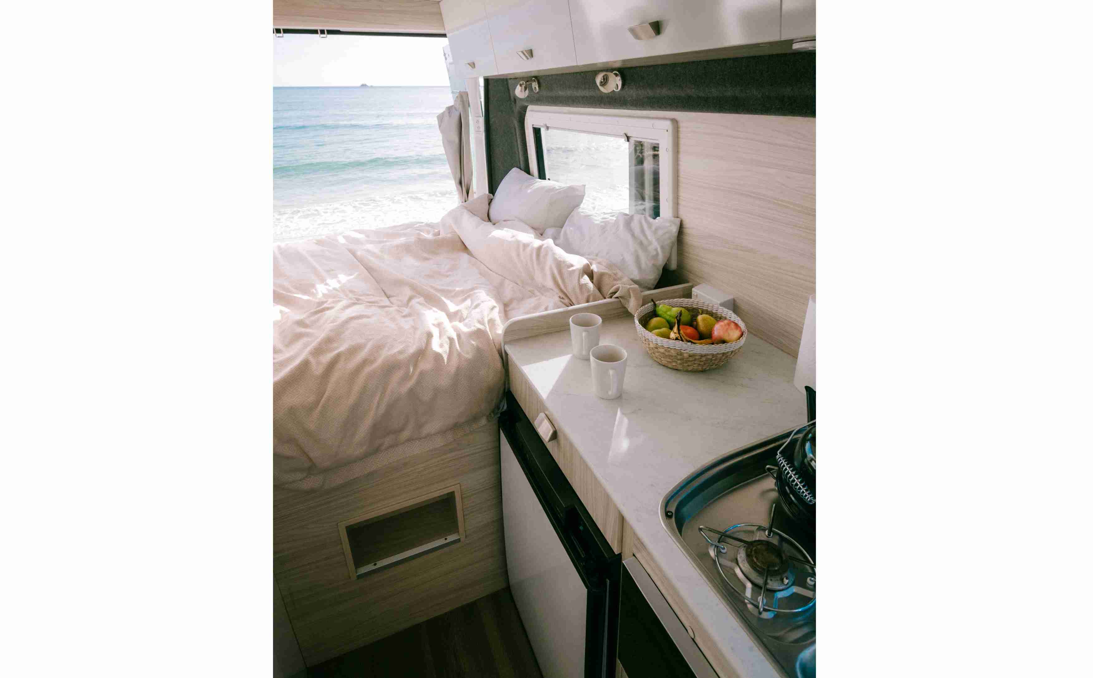 Campervan interior showing a bed facing the ocean, next to a kitchen counter with mugs and a fruit bowl.