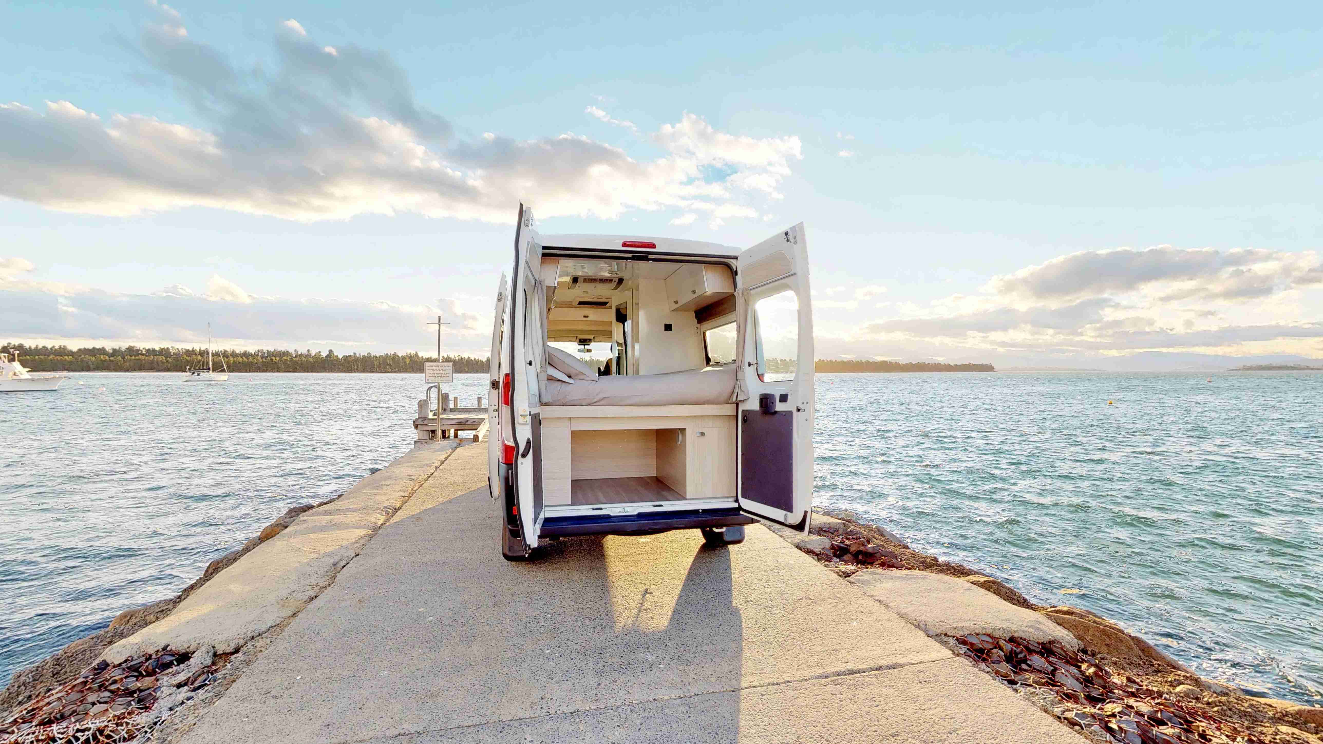 White campervan with open rear doors parked on a concrete jetty, showing a bed and storage space overlooking the sea.
