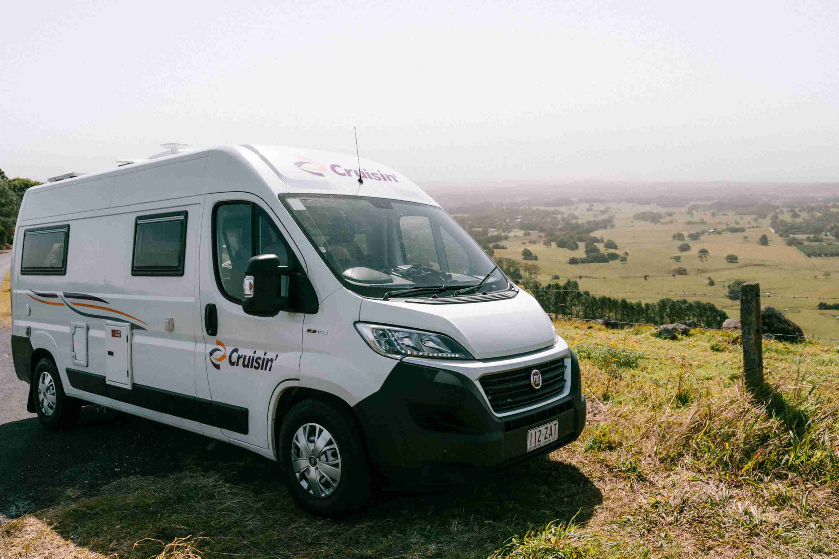 White Cruisin' campervan parked on a grassy hillside overlooking a vast green valley.