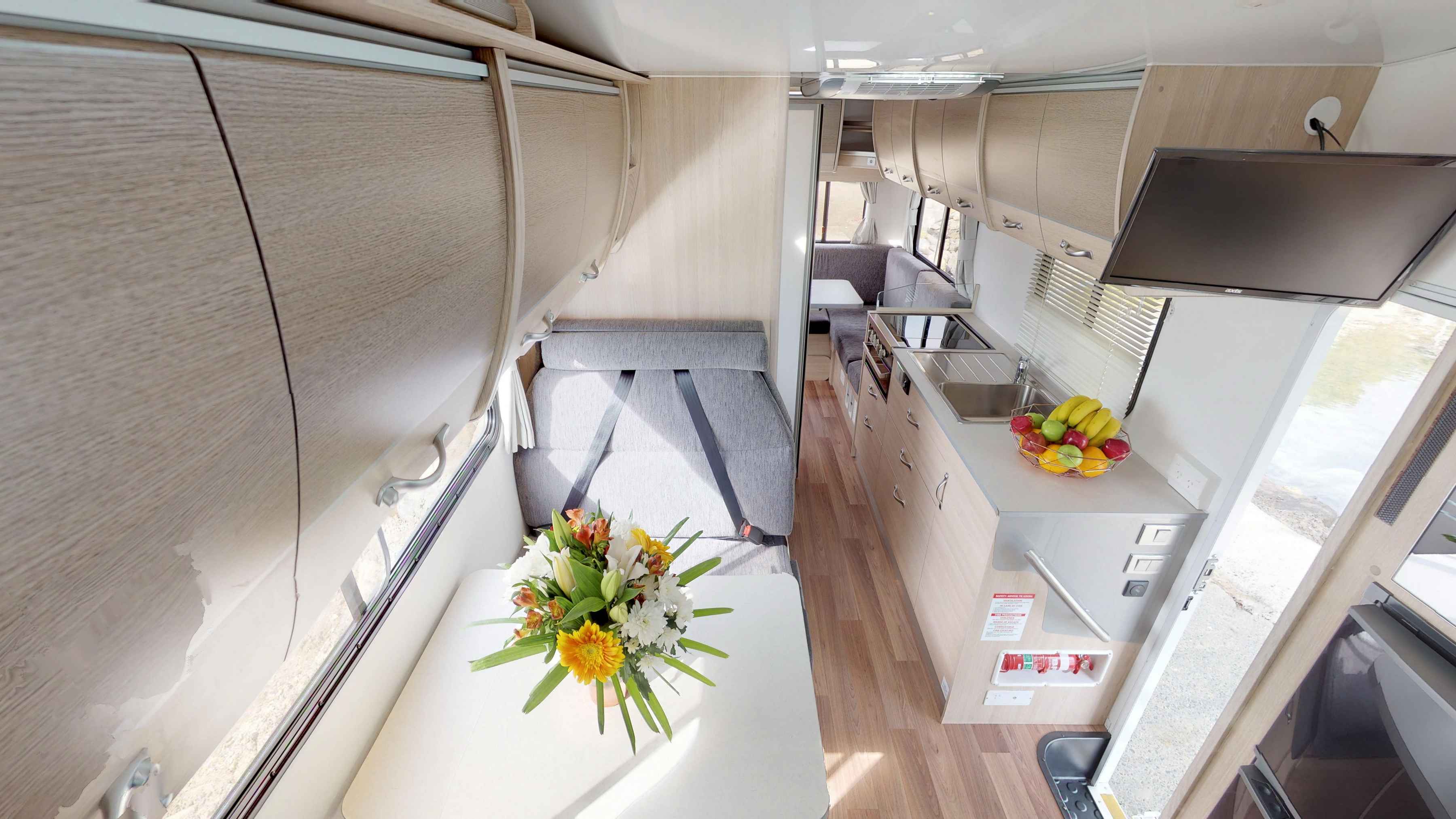 Modern campervan interior featuring light timber cabinetry, grey dinette seating, a table, and a compact kitchen.