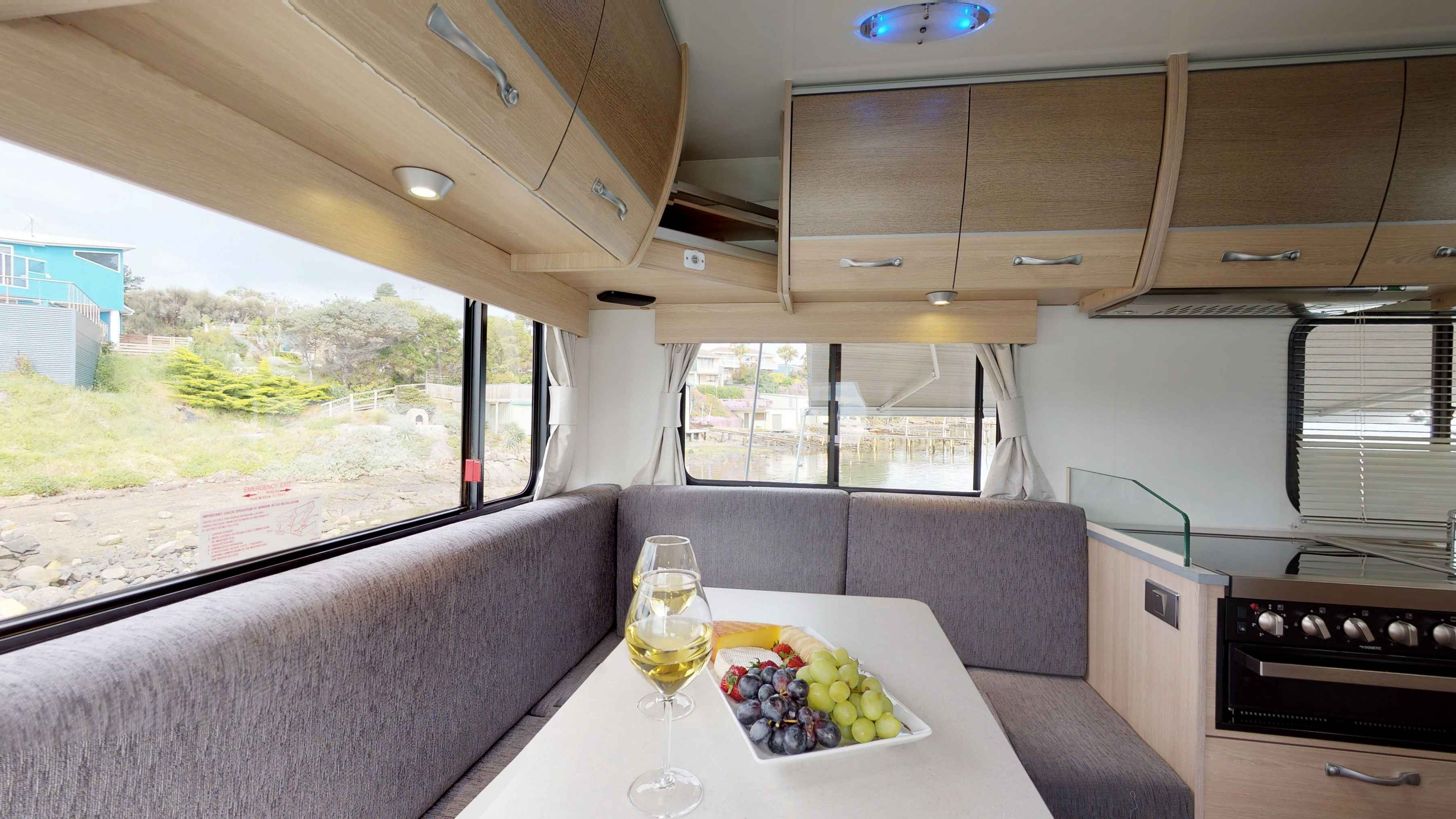 Campervan interior featuring a grey fabric dining booth, wine and fruit platter, and timber overhead cabinets.