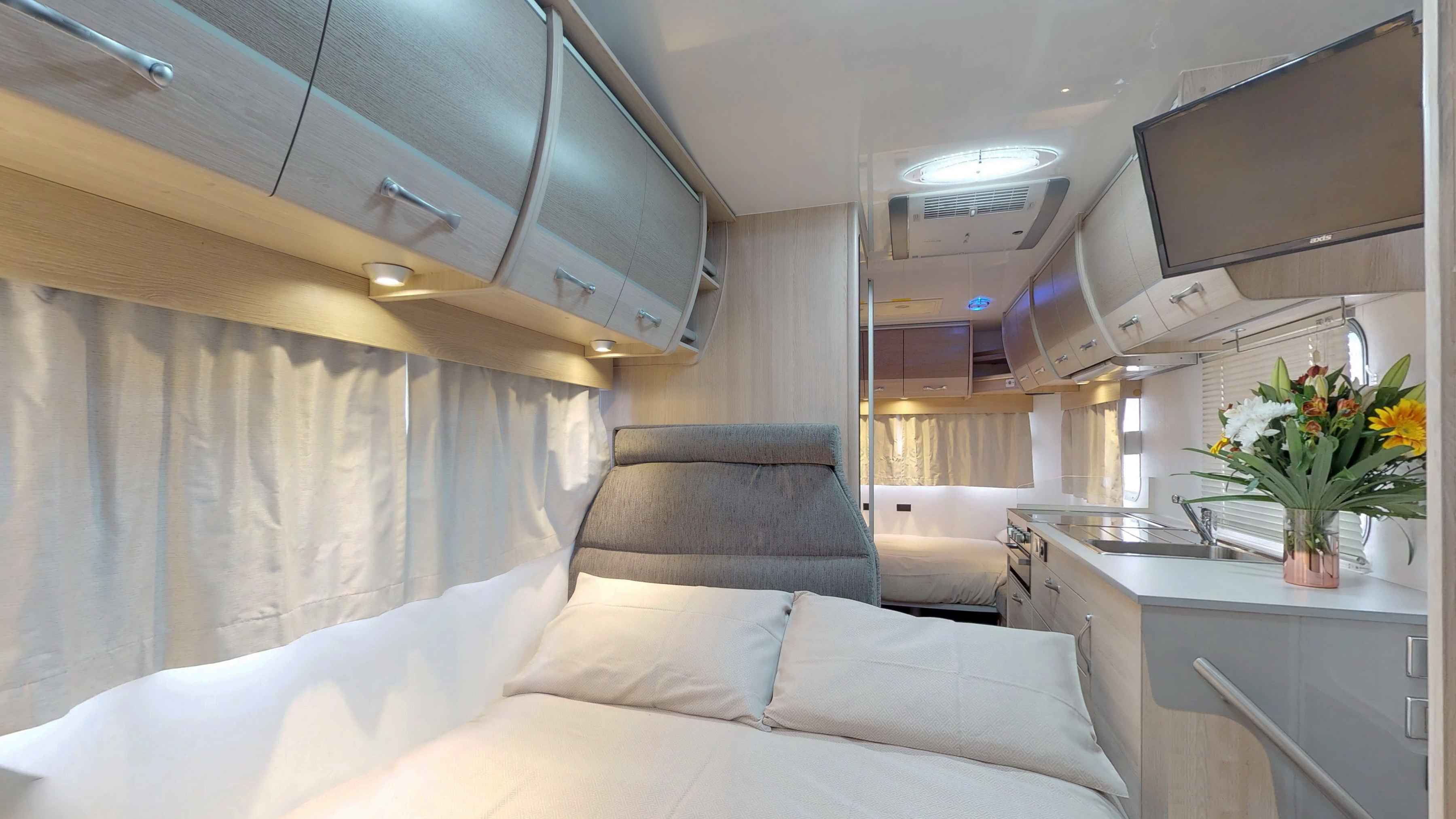 Campervan interior featuring a double bed, overhead cabinets, and a compact kitchenette with a wall-mounted TV.