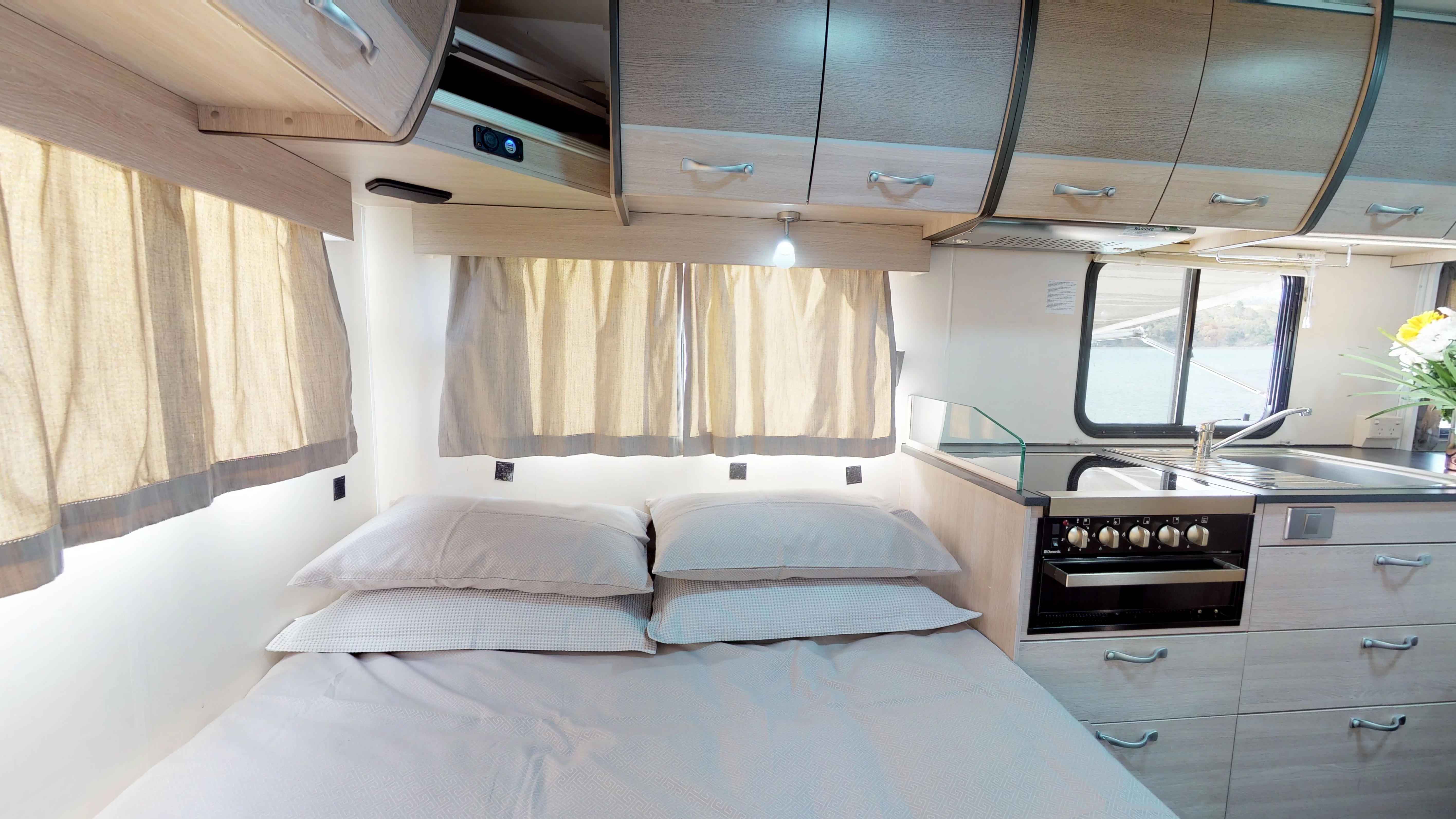 Campervan interior featuring a double bed with grey pillows next to a compact kitchen stove, oven, and cabinets.