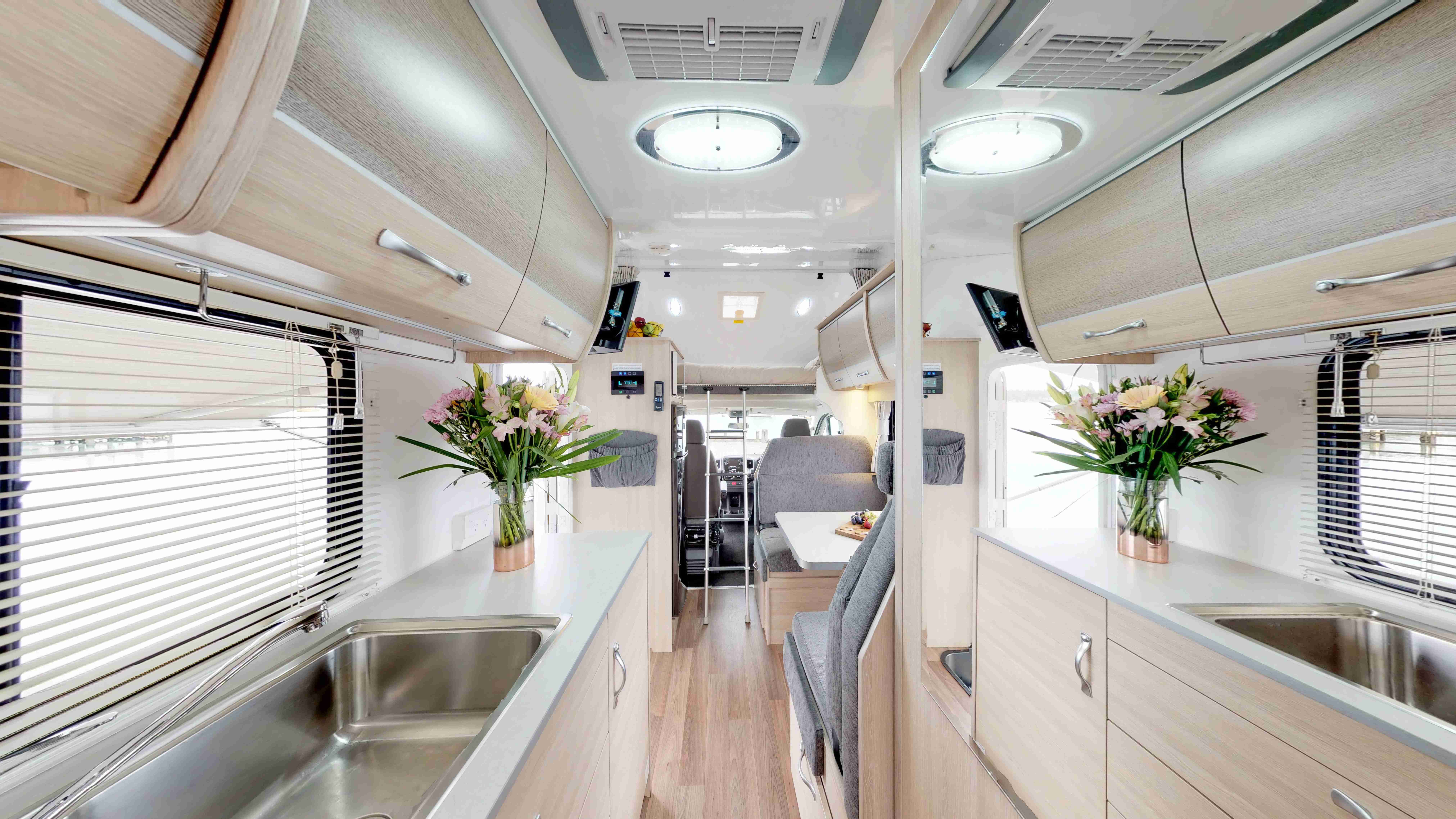 Interior of a modern campervan with light timber-look cabinetry, a stainless steel sink, and grey upholstered seating.