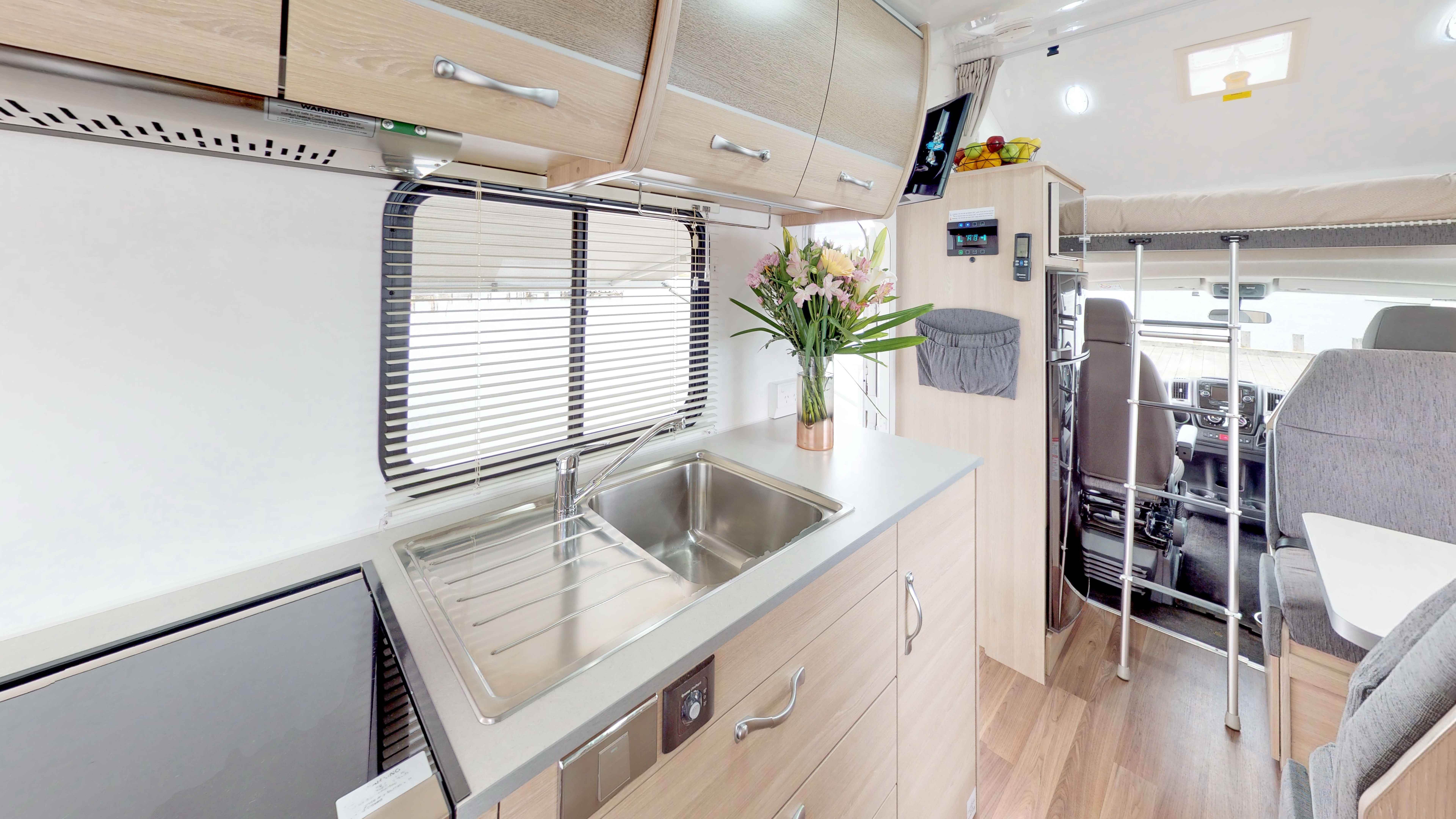 Campervan interior featuring a stainless steel sink, light timber cabinets, and a ladder to the over-cab bed.