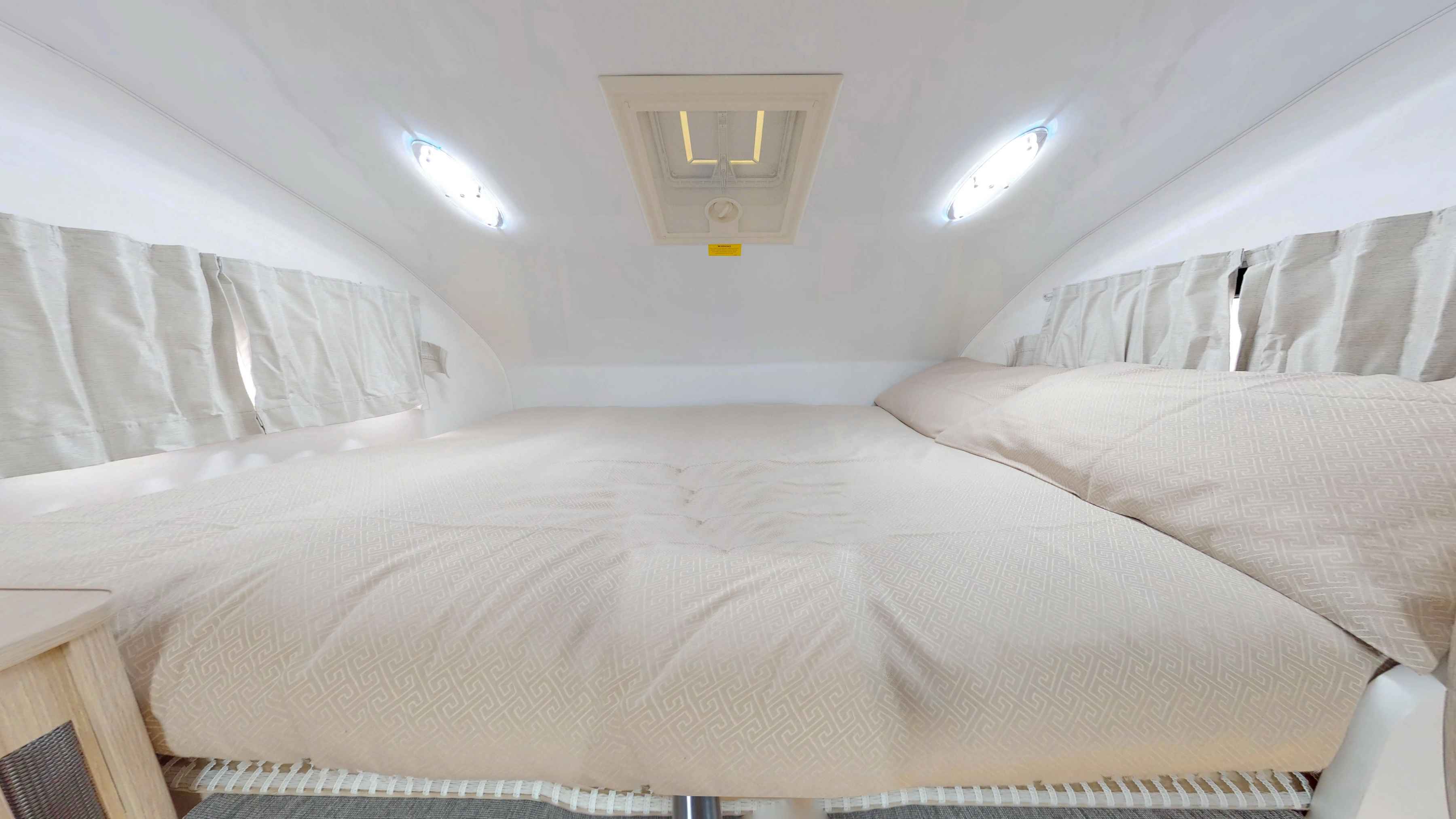 Campervan loft double bed with patterned beige bedding, side curtains, dual LED lights, and a central ceiling hatch.