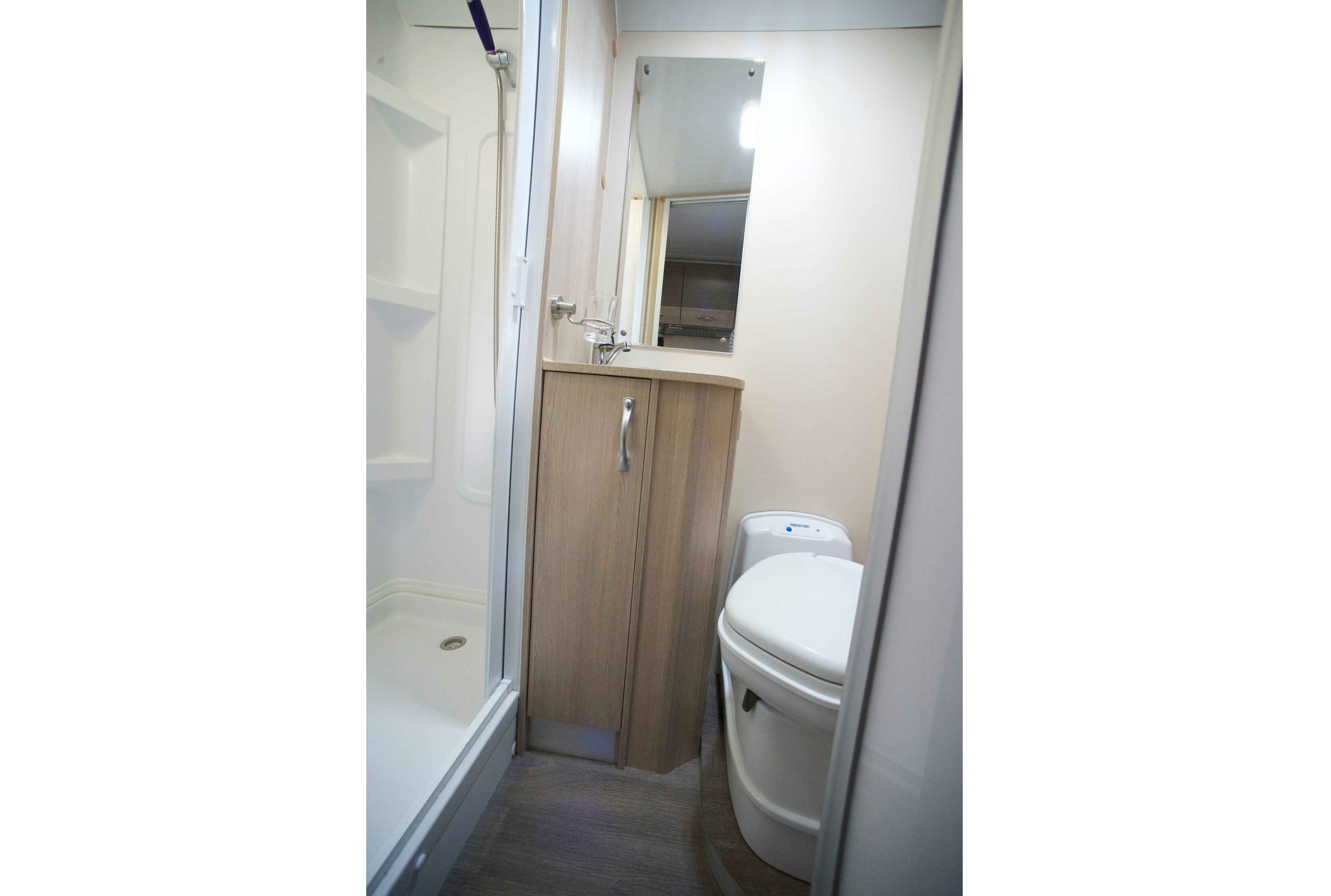 Compact campervan bathroom featuring a white shower cubicle, light timber vanity with a mirror, and a cassette toilet.