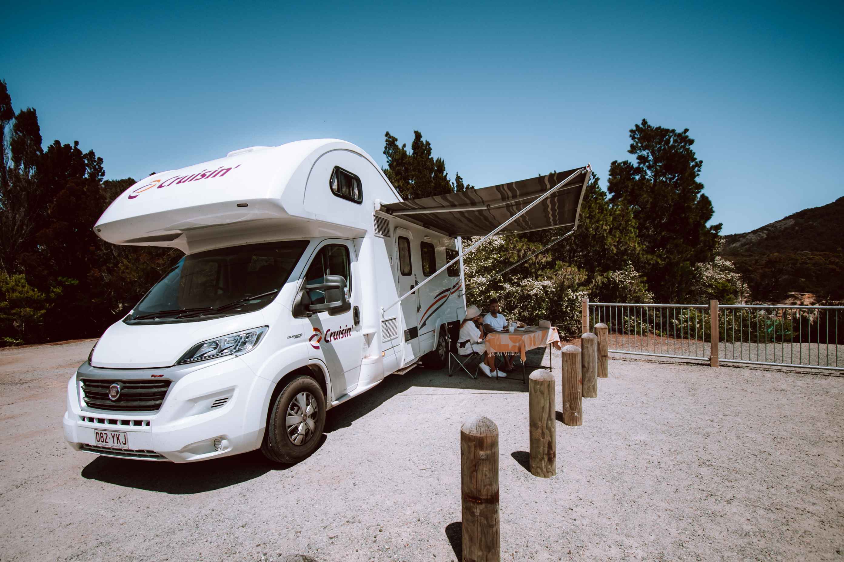 White Cruisin' campervan with an extended awning and two people sitting at an outdoor table, parked on gravel.