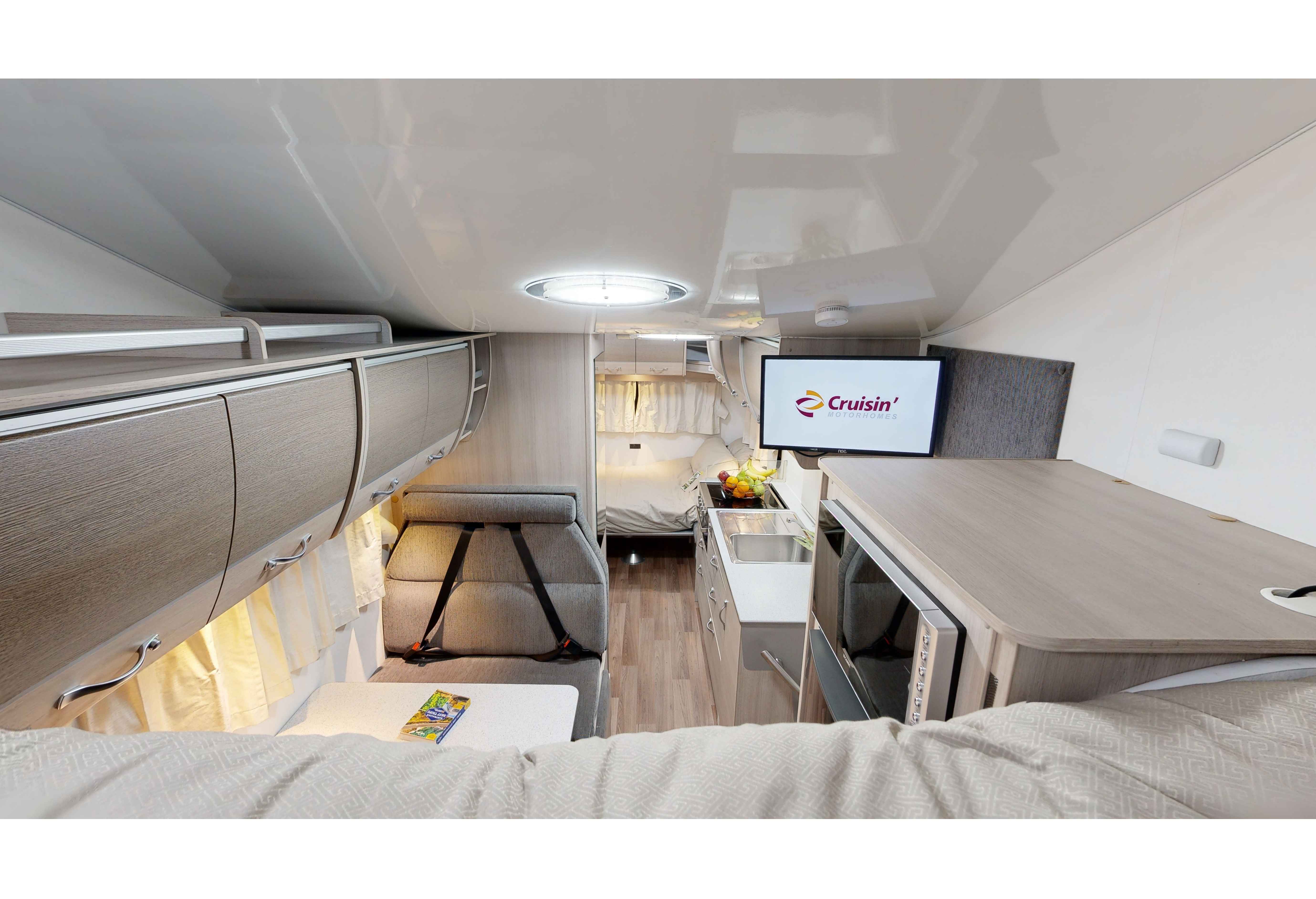 Interior view of a modern campervan with grey seating, compact kitchen with a TV, and a rear sleeping area.