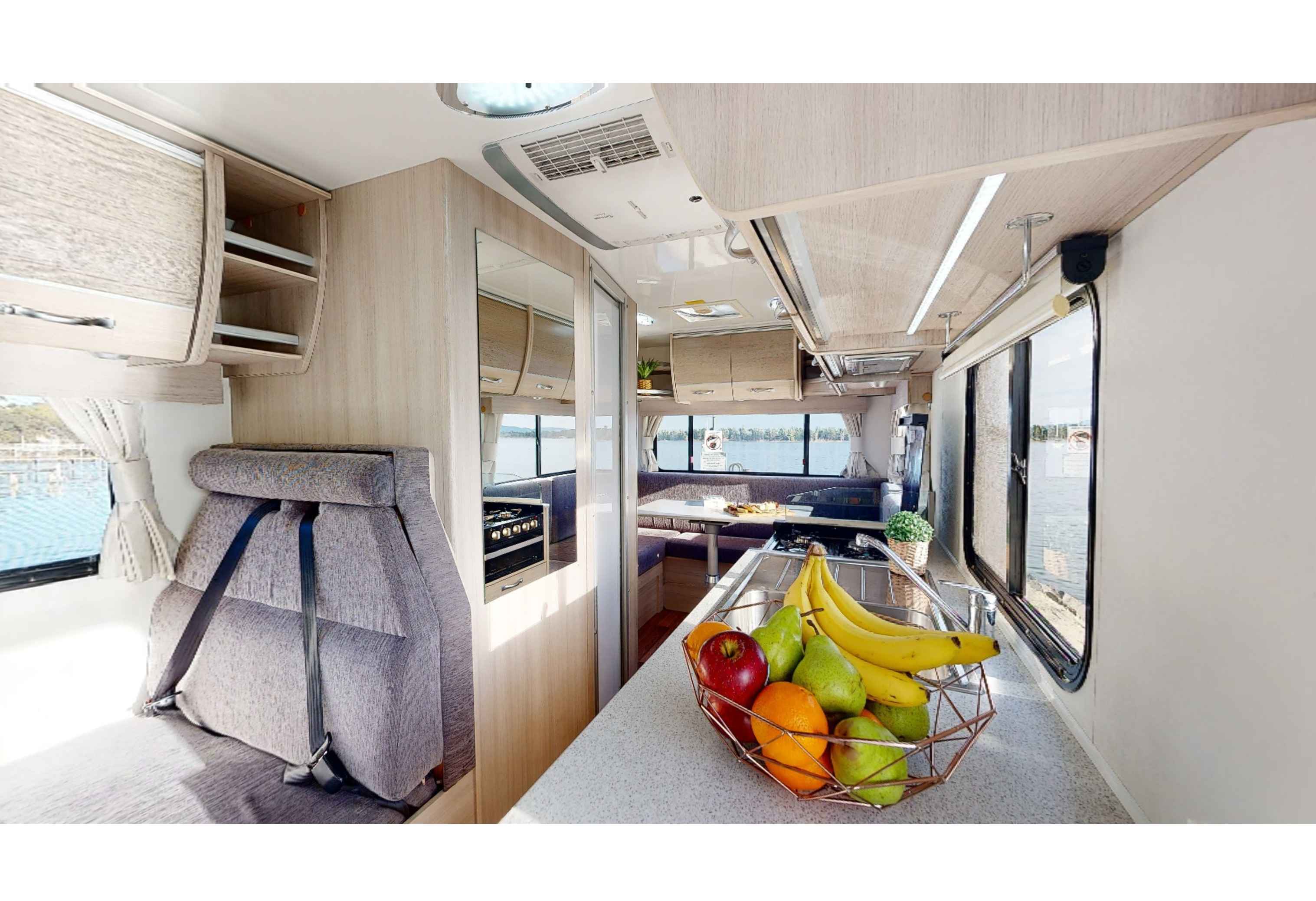 Campervan interior showing a grey seat, kitchen counter with a fruit bowl, and rear dining area overlooking the water.
