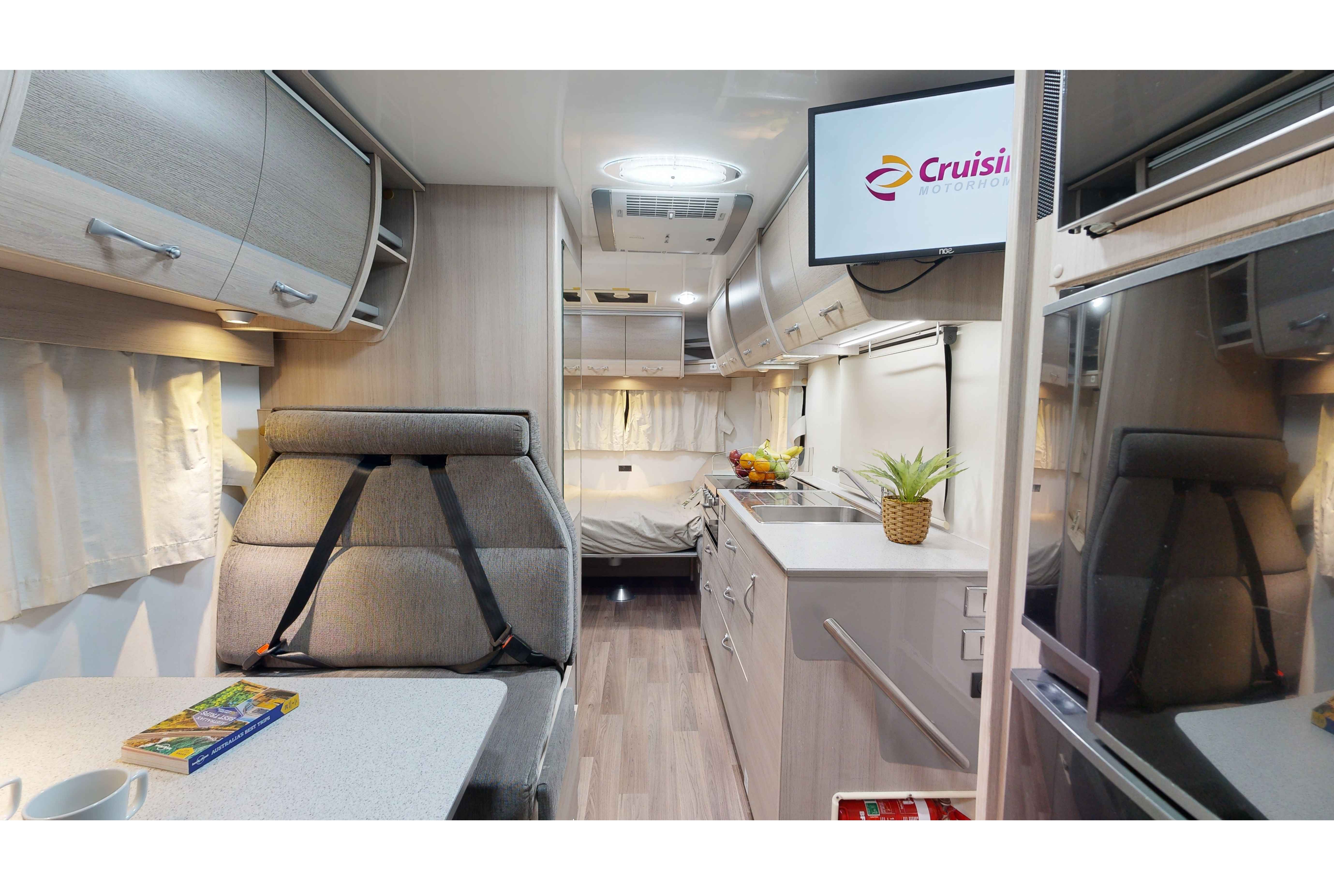 Interior of a modern campervan with grey dining booth, compact kitchen, overhead TV, and a bed in the background.