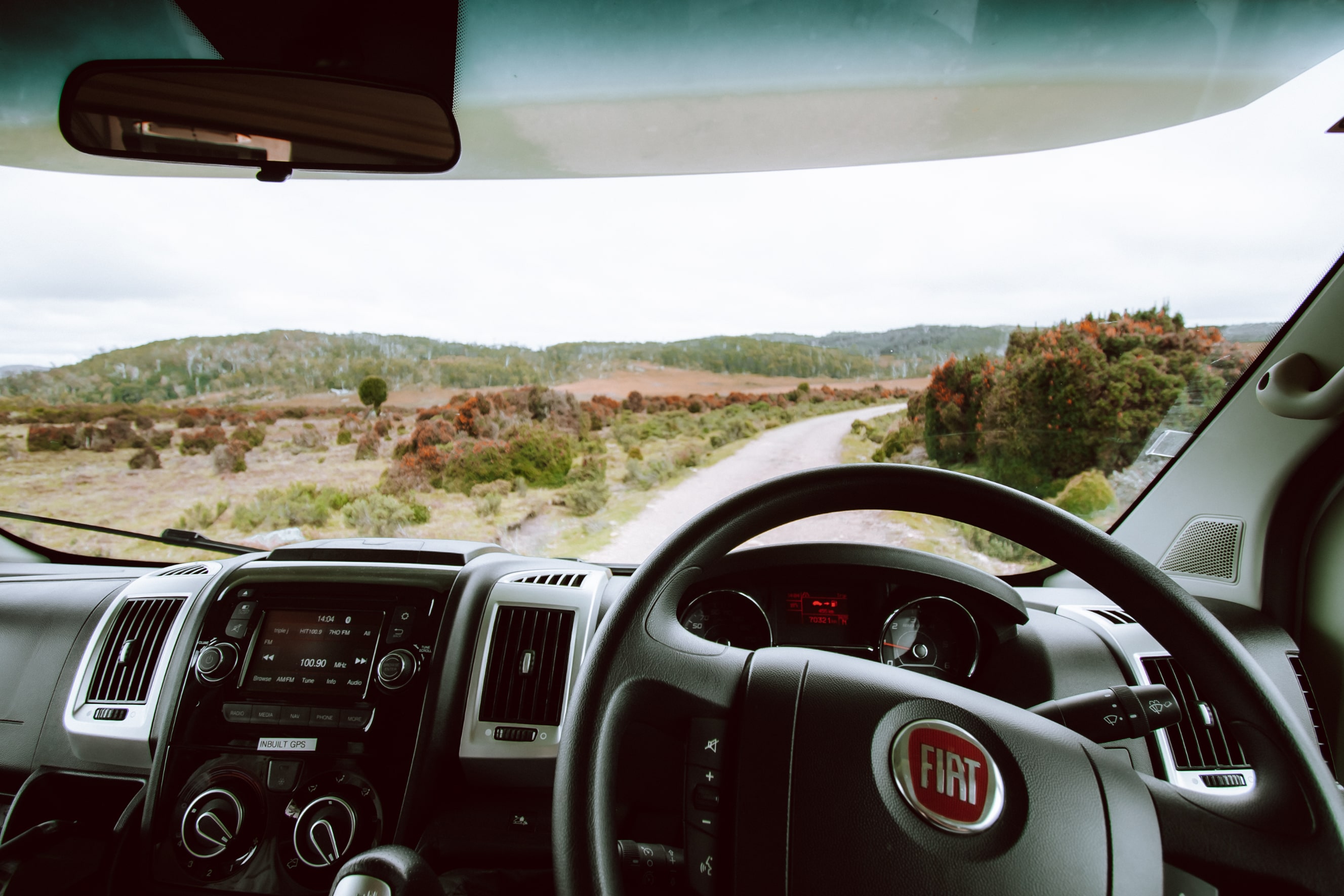 View from the driver's seat of a Fiat campervan looking out onto a winding dirt road through a rural landscape.