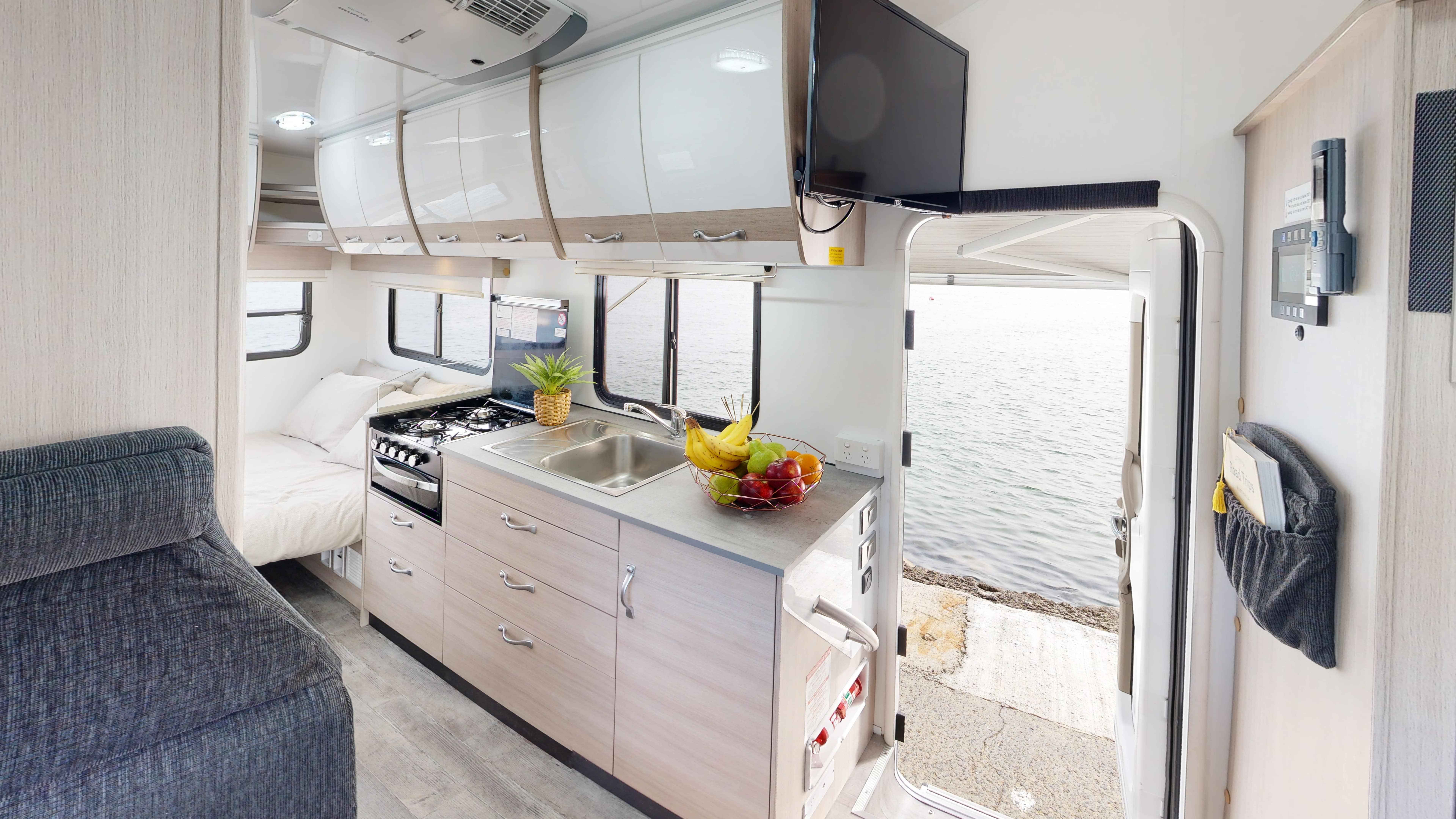 Interior of a campervan showing a kitchenette, bed, and open door looking directly onto water.