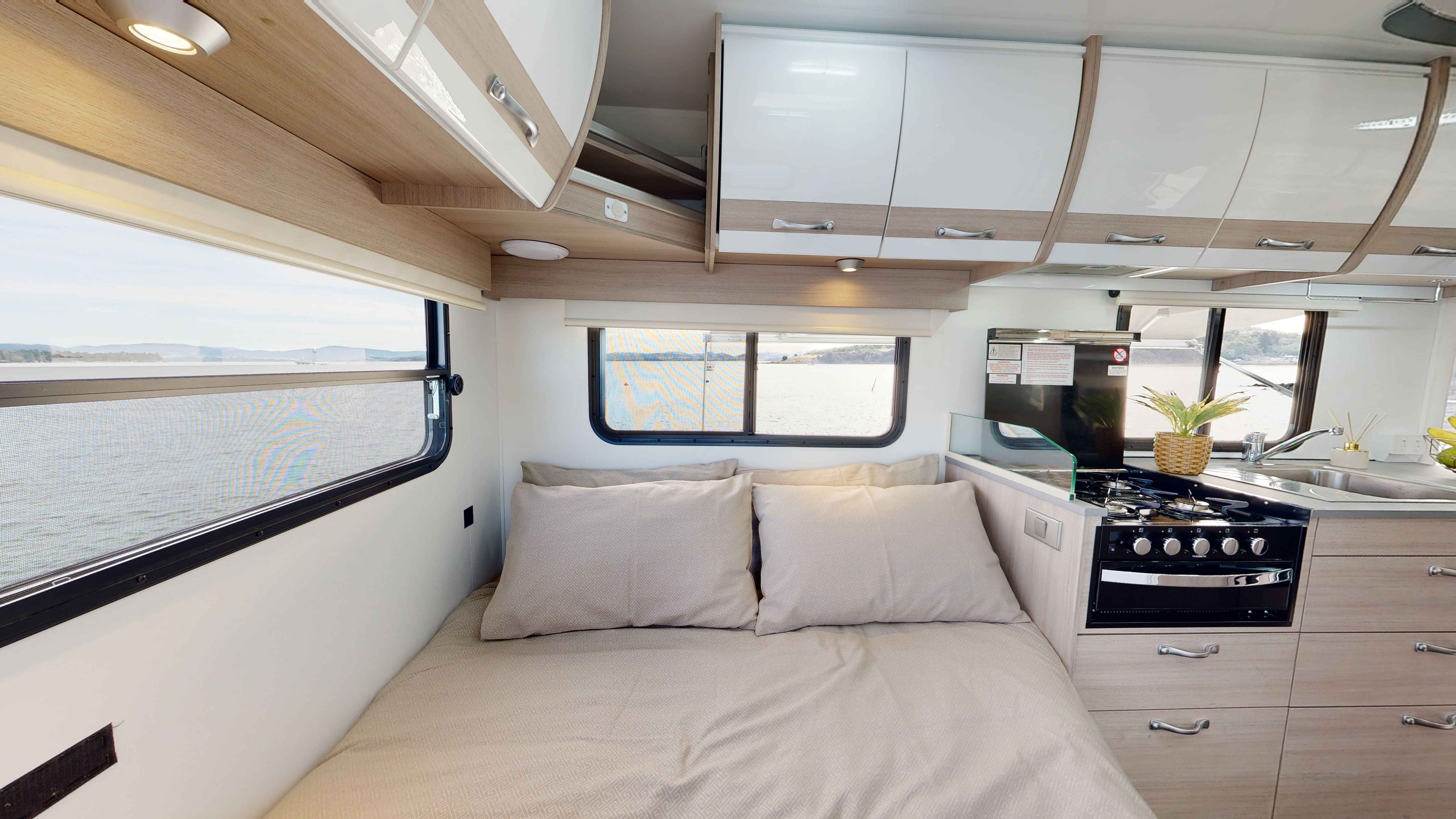 Modern campervan interior featuring a double bed, compact kitchen with stove, and windows overlooking water.