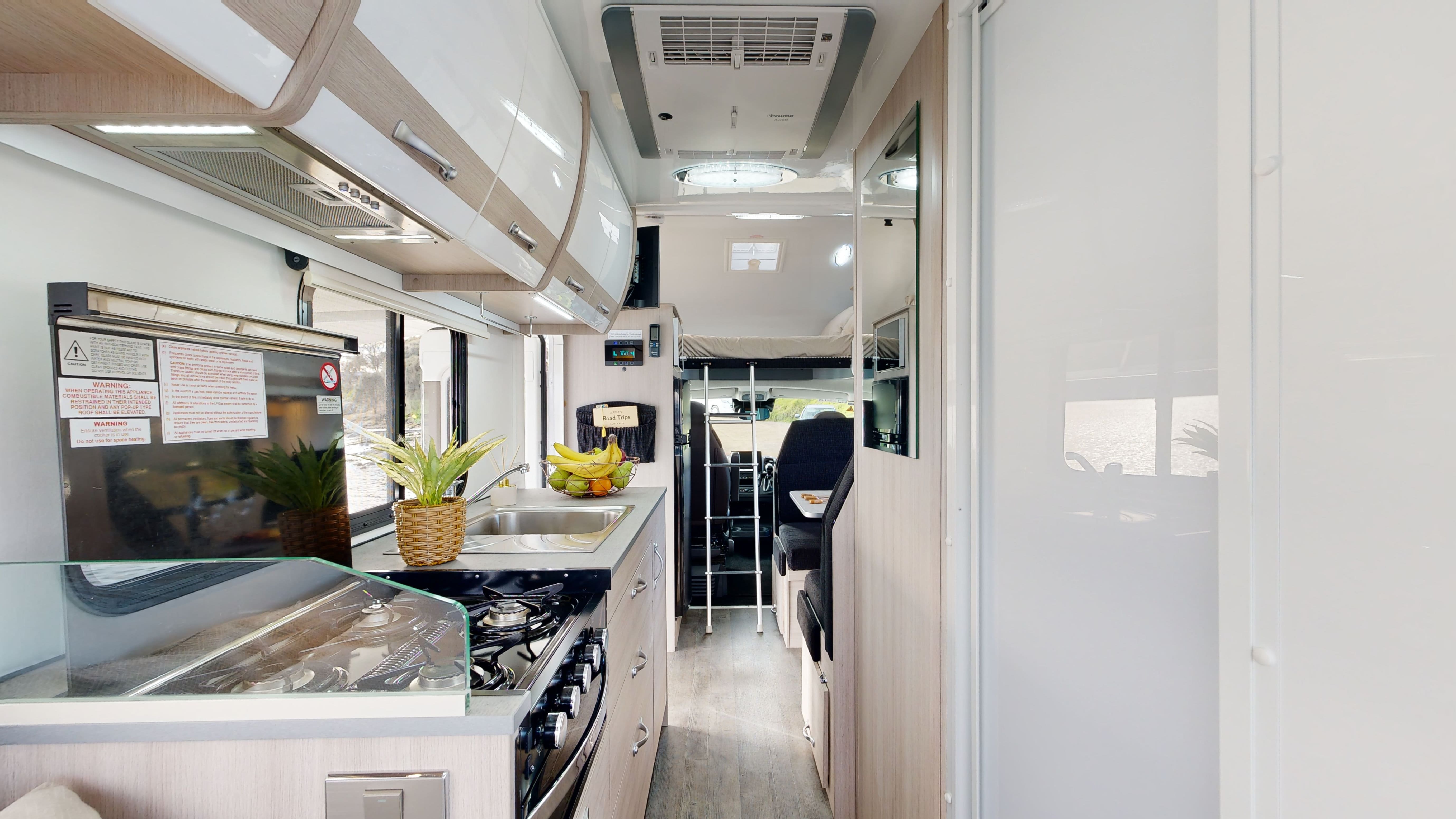 Modern campervan interior featuring a kitchen with a stove, sink, white cabinets, and an aisle leading to a loft bed.